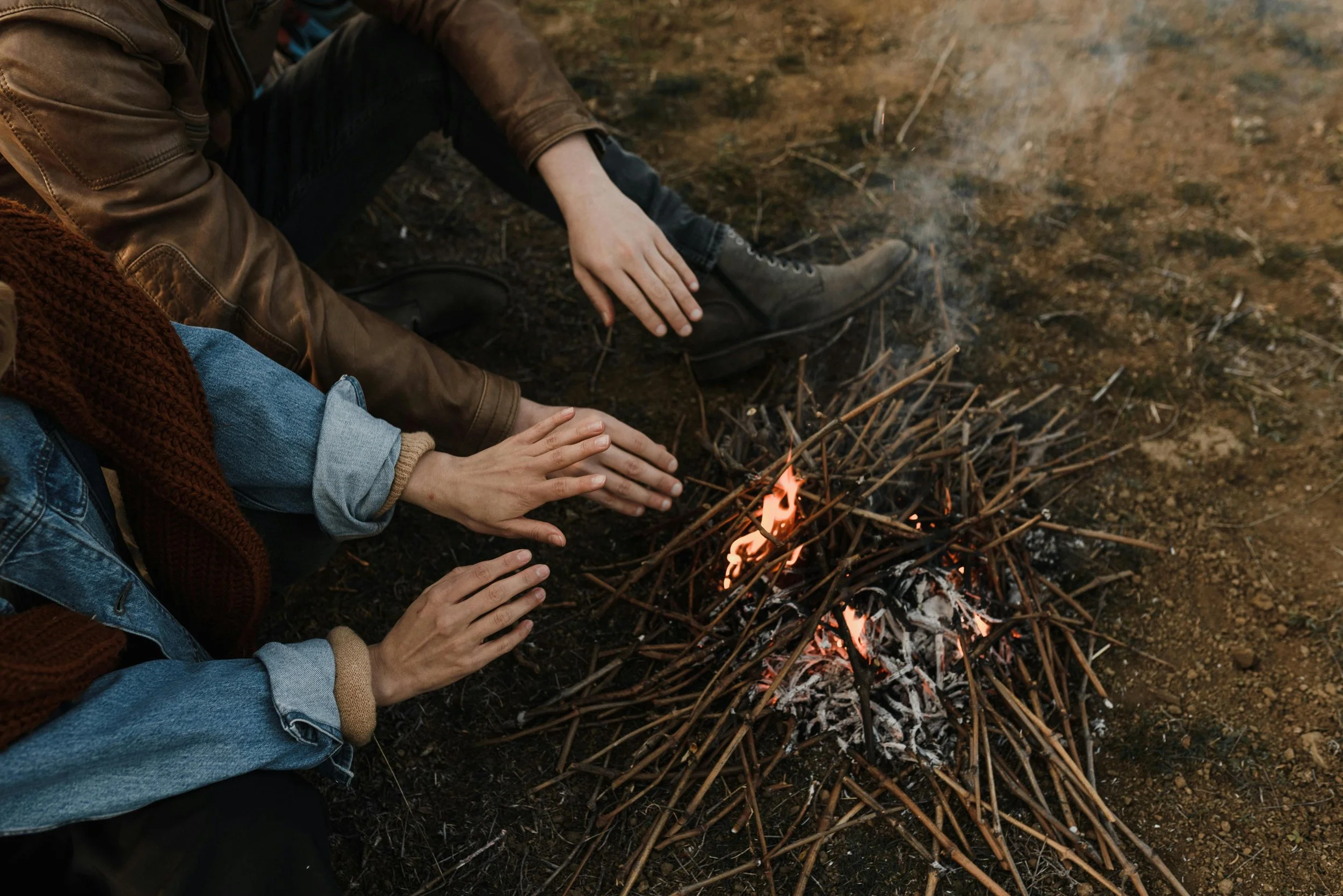 Two people wearing casual clothes and boots warming their hands over a small campfire made of sticks outdoors on dirt ground.
