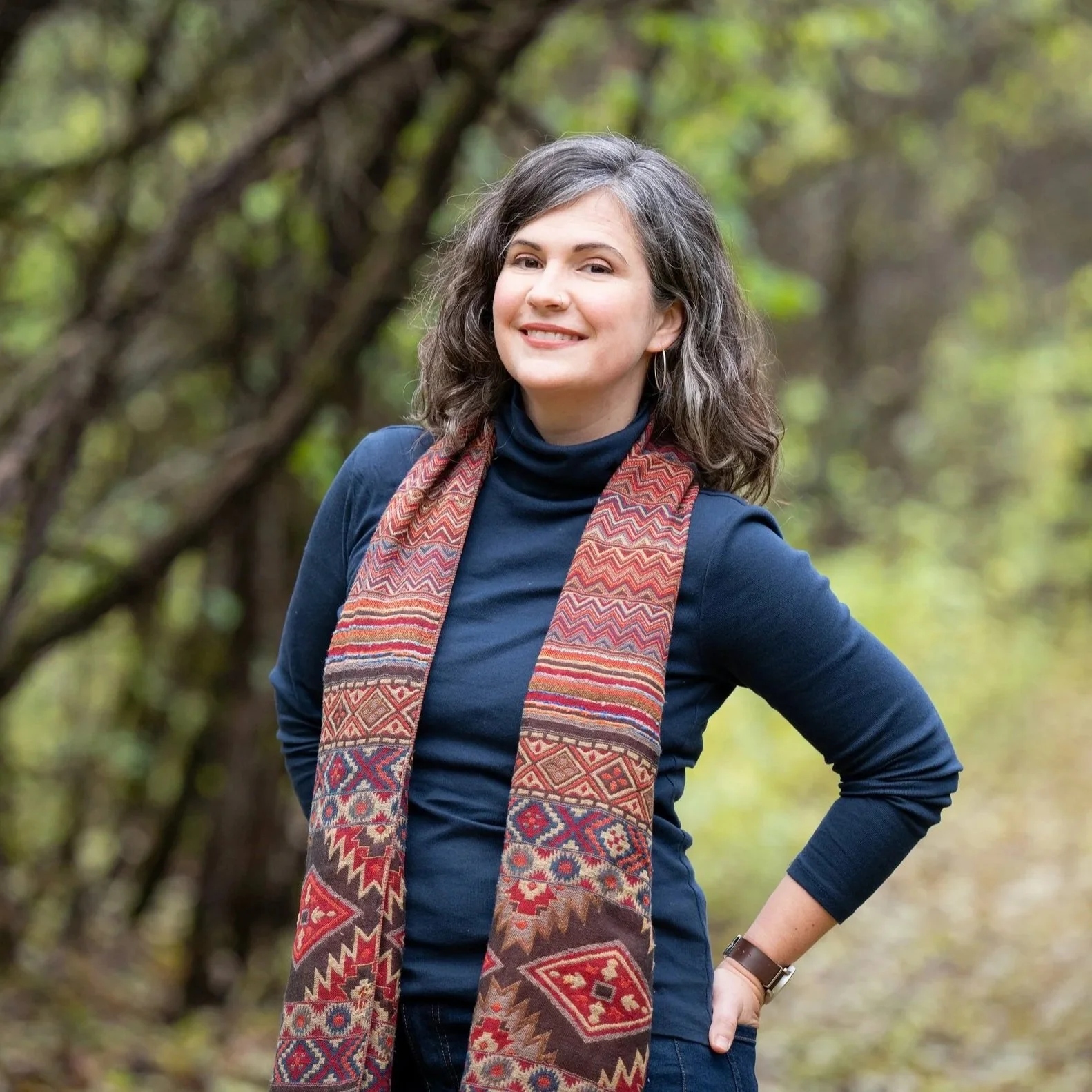 Elodie Mantha, twice-certified intergrative changework coach stands outdoors in a forest, wearing a navy blue turtleneck and a colorful patterned scarf, looking at the camera and smiling with her hand on her hip.