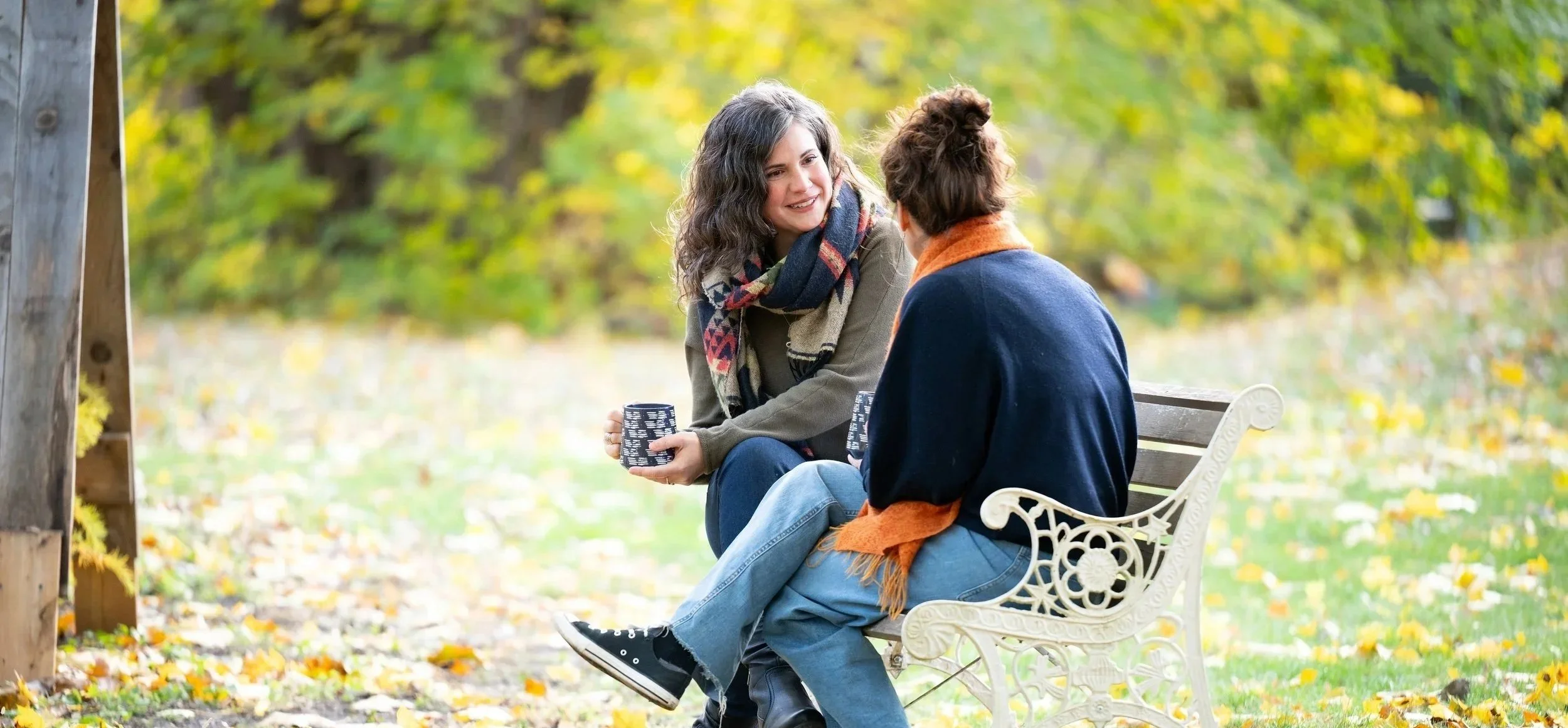Elodie Mantha sits on a park bench with a client, engaged in a coaching conversation amidst yellow and green autumn leaves. Both women hold blue coffee mugs in their hands, and are smiling.