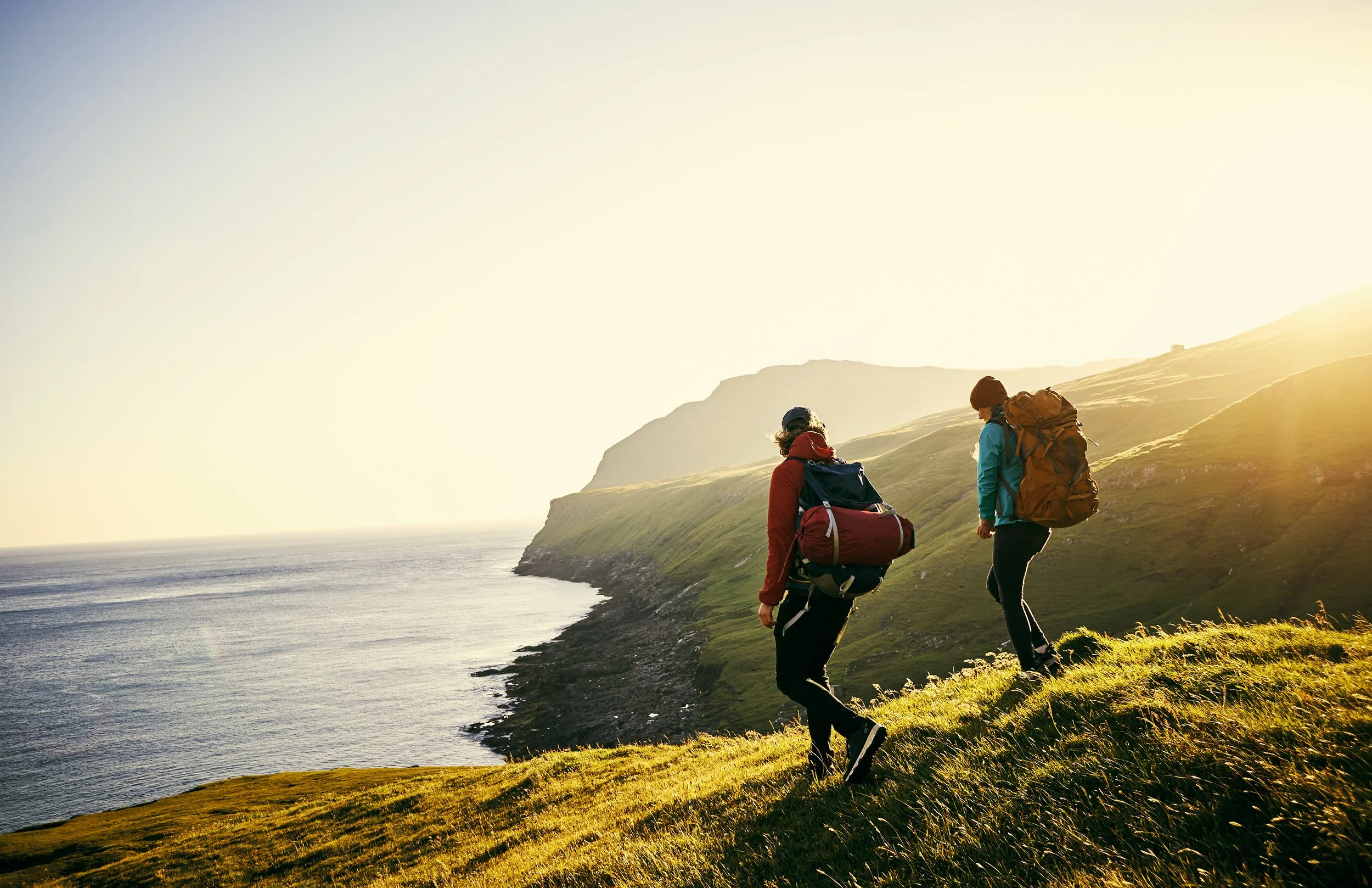 Two hikers with backpacks walking along a grassy hill overlooking the ocean at sunrise or sunset.