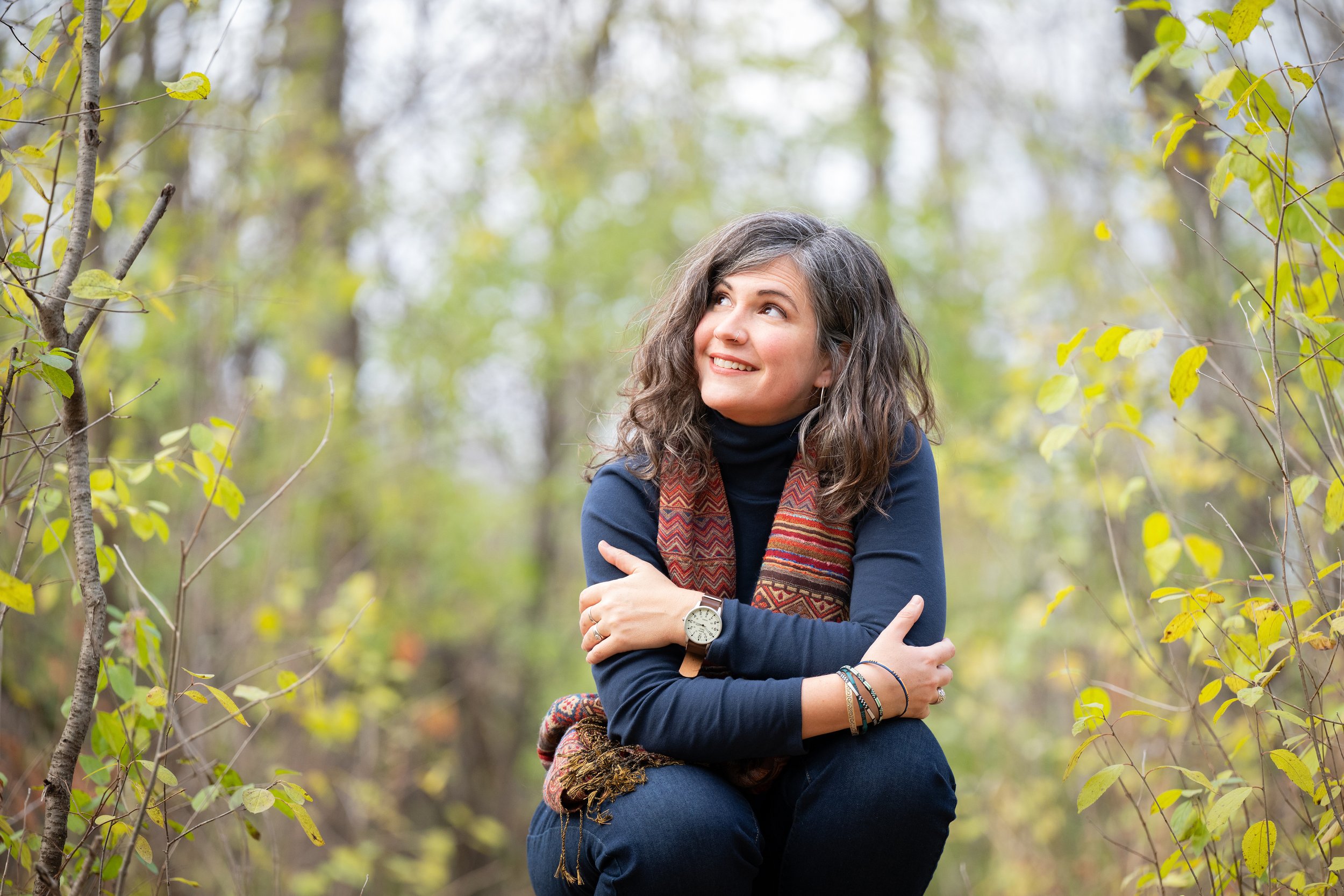 Elodie Mantha, twice-certified intergrative changework coach,  is sitting outdoors among trees with yellow and green leaves, smiling and looking upward, wearing a navy blue turtleneck, a patterned scarf, and a watch.
