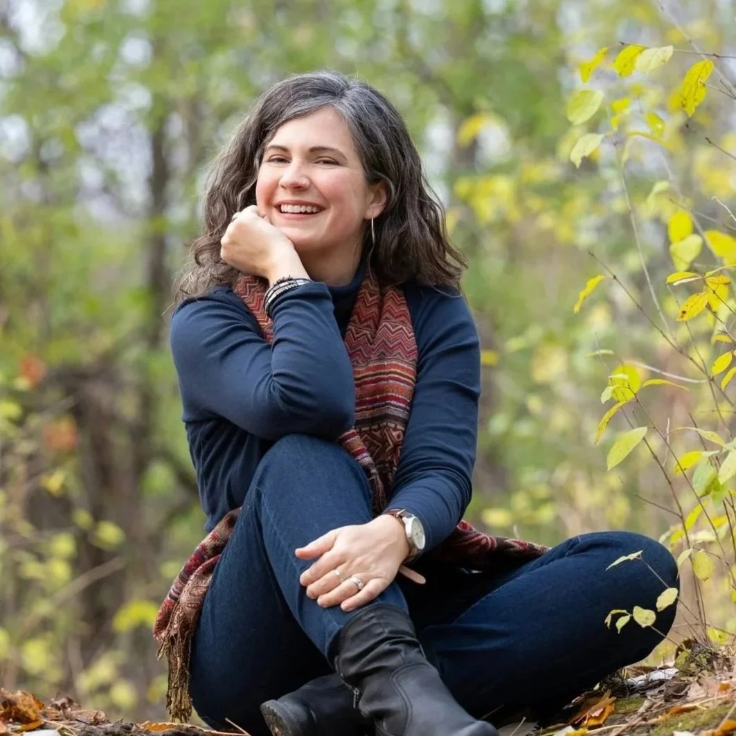 Elodie Mantha is sitting outdoors in a forest, looking at the camera and smiling. She has curly brown hair and is dressed in a dark blue long-sleeve shirt, dark blue jeans, and black boots, with a multicolored scarf and watch.