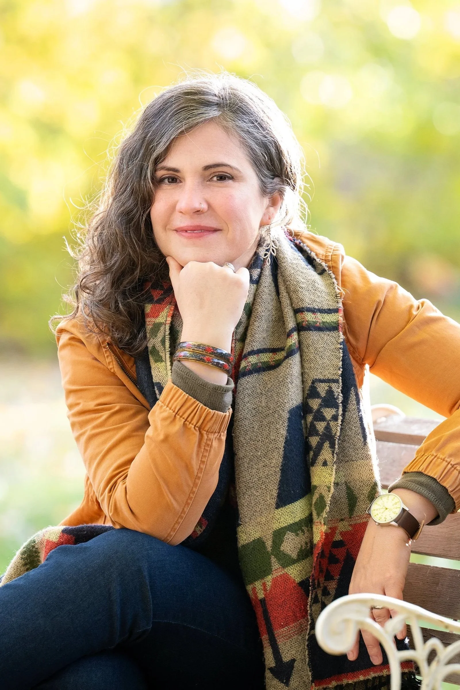 Elodie Mantha, twice-certified intergrative changework, with long wavy dark hair sits outdoors on a bench, resting her chin on her hand, smiling slightly, with a patterned scarf, a tan jacket, and a watch, in front of a blurred autumnal background.