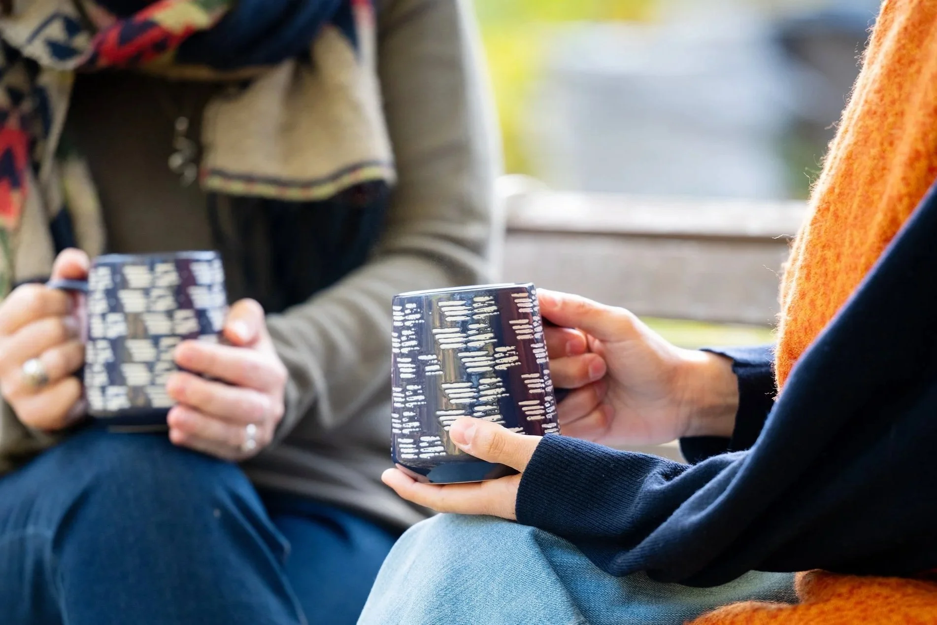 Two people sitting outdoors, each holding a matching ceramic mug with a dark background and white vertical lines, having a deeply engaging coaching conversation.