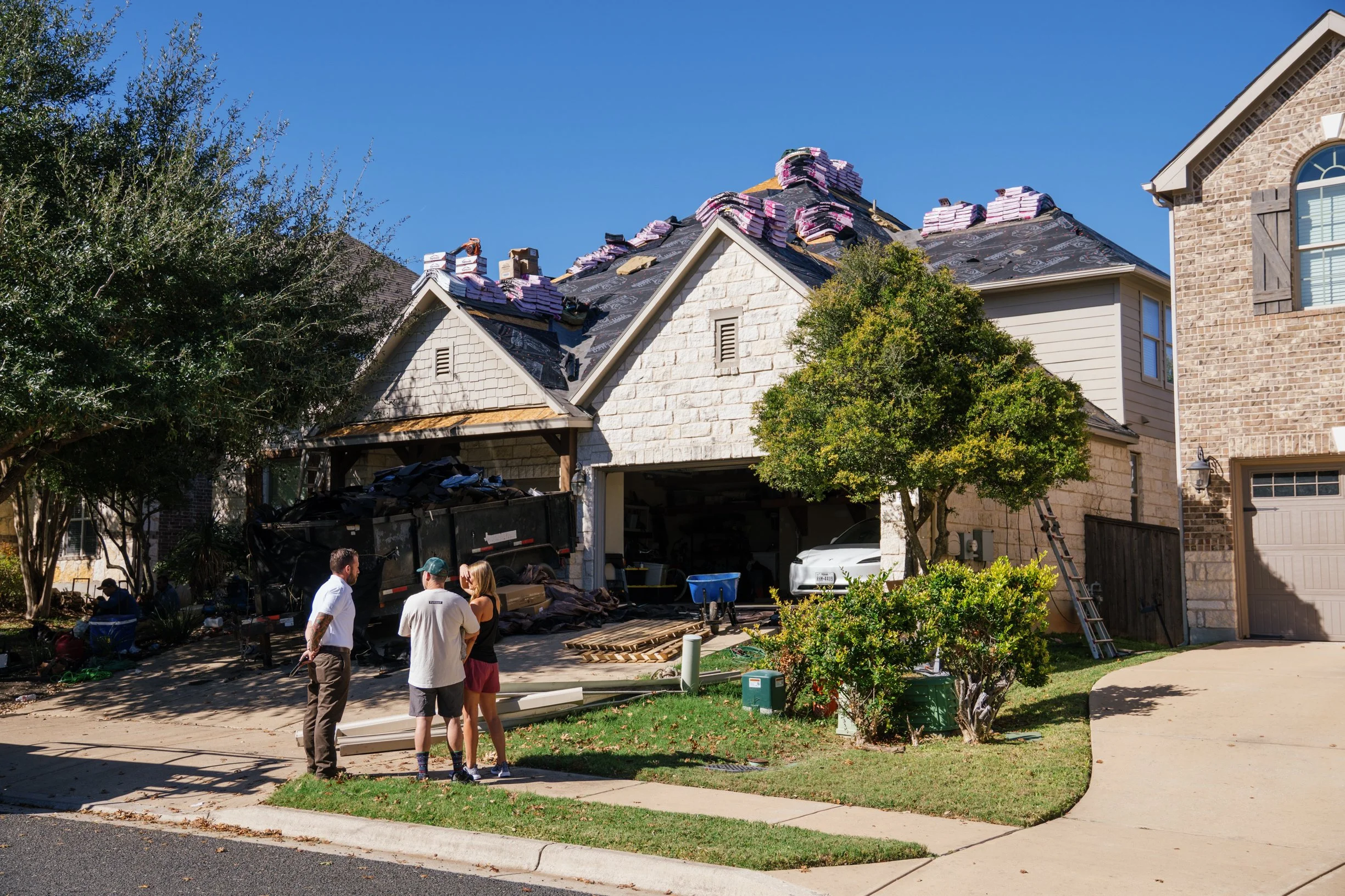 People standing in front of a house under renovation, with roofing materials and construction workers present.
