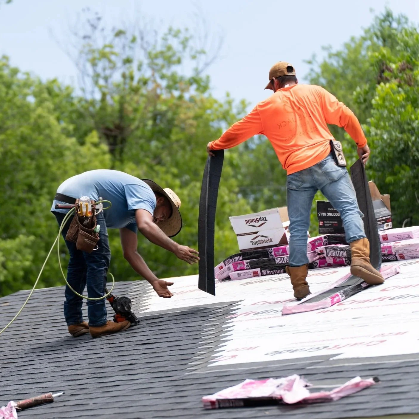 Two workers installing roofing materials on a house roof outdoors, surrounded by green trees.
