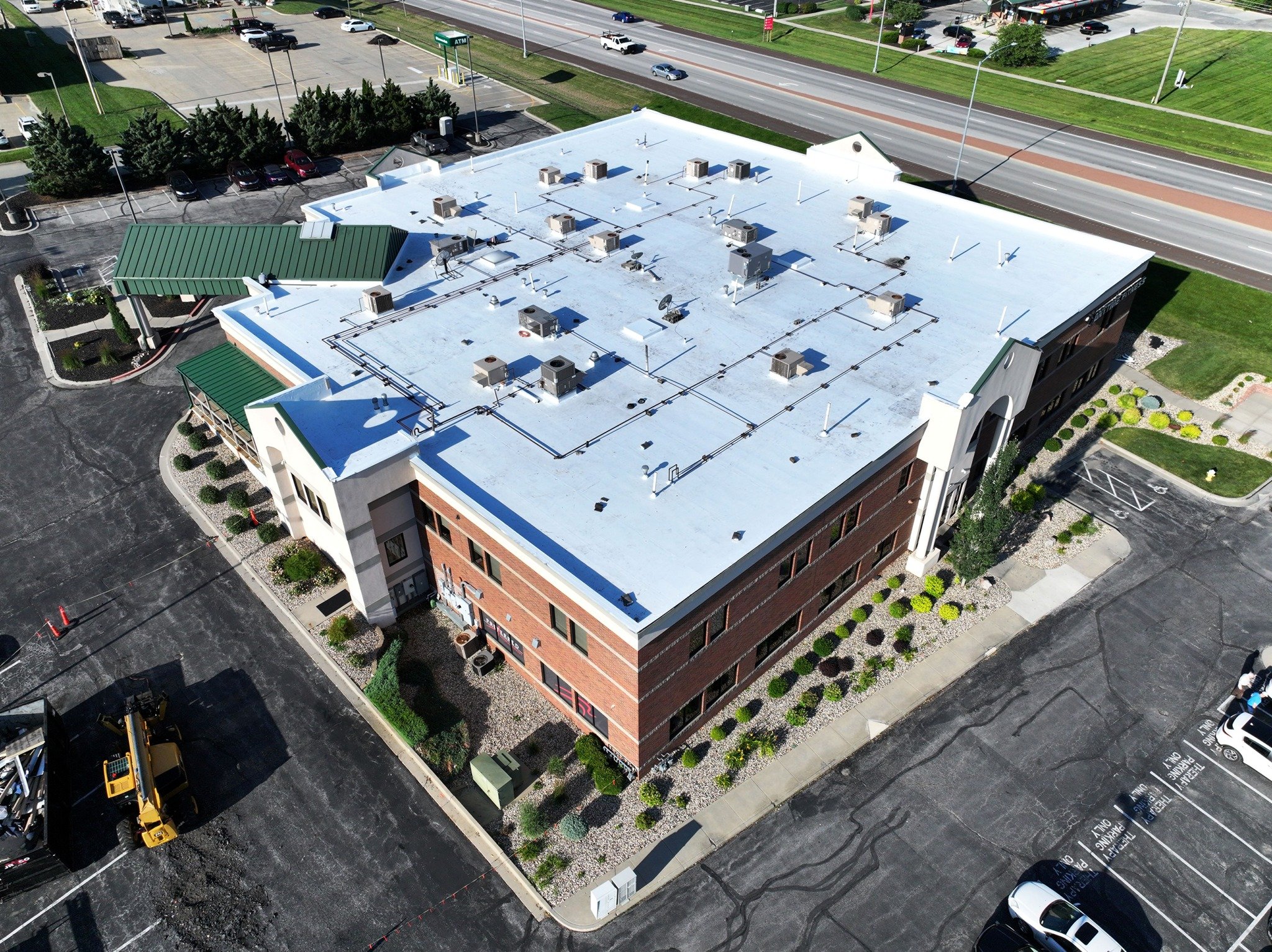 Aerial view of a multi-story commercial building with a flat roof, rooftop HVAC units, and a parking lot with some cars. The building is adjacent to a busy highway with multiple lanes of traffic and green landscaped areas.
