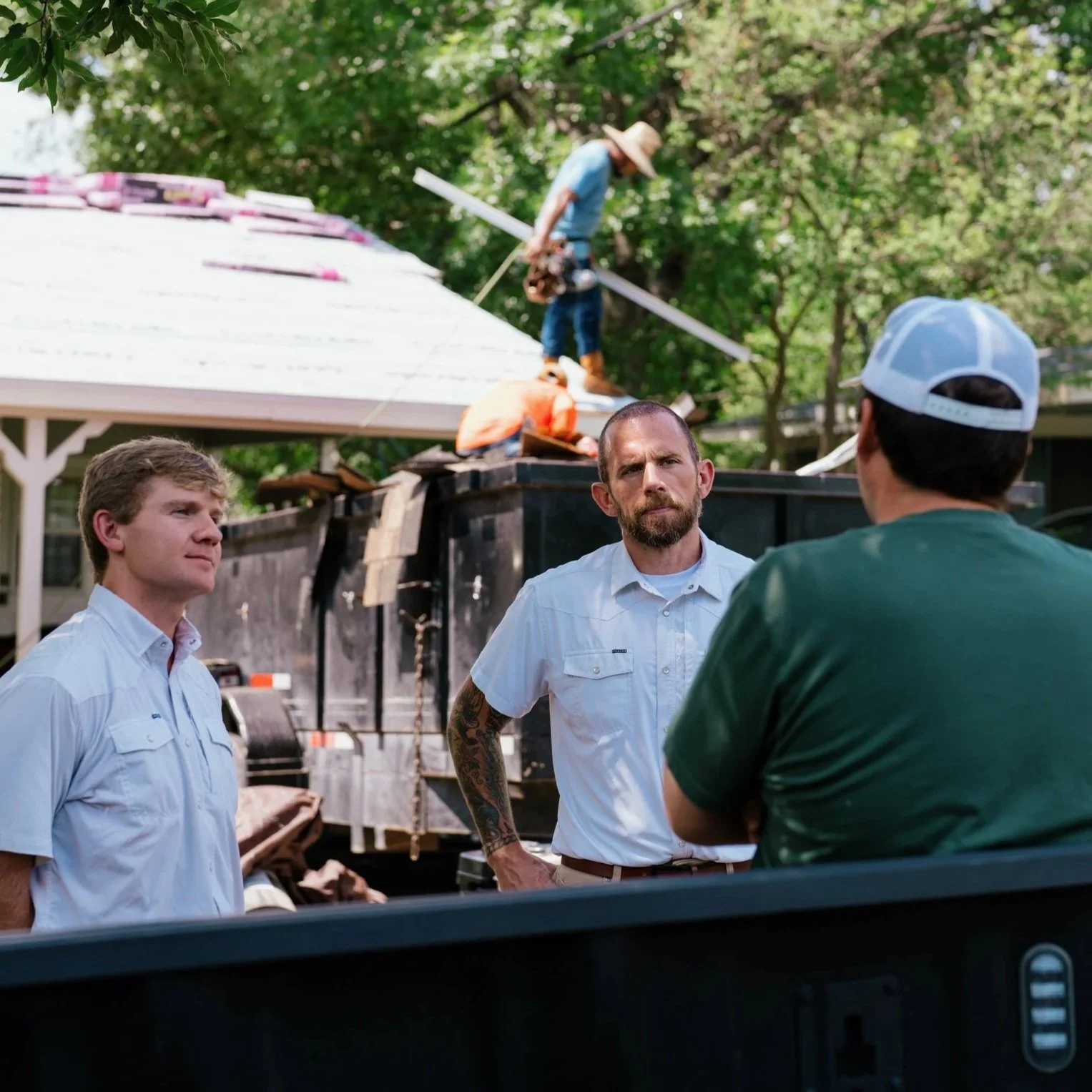 Three men engaged in conversation outdoors near a pickup truck with construction workers on the roof installing or repairing a roof, surrounded by green trees.