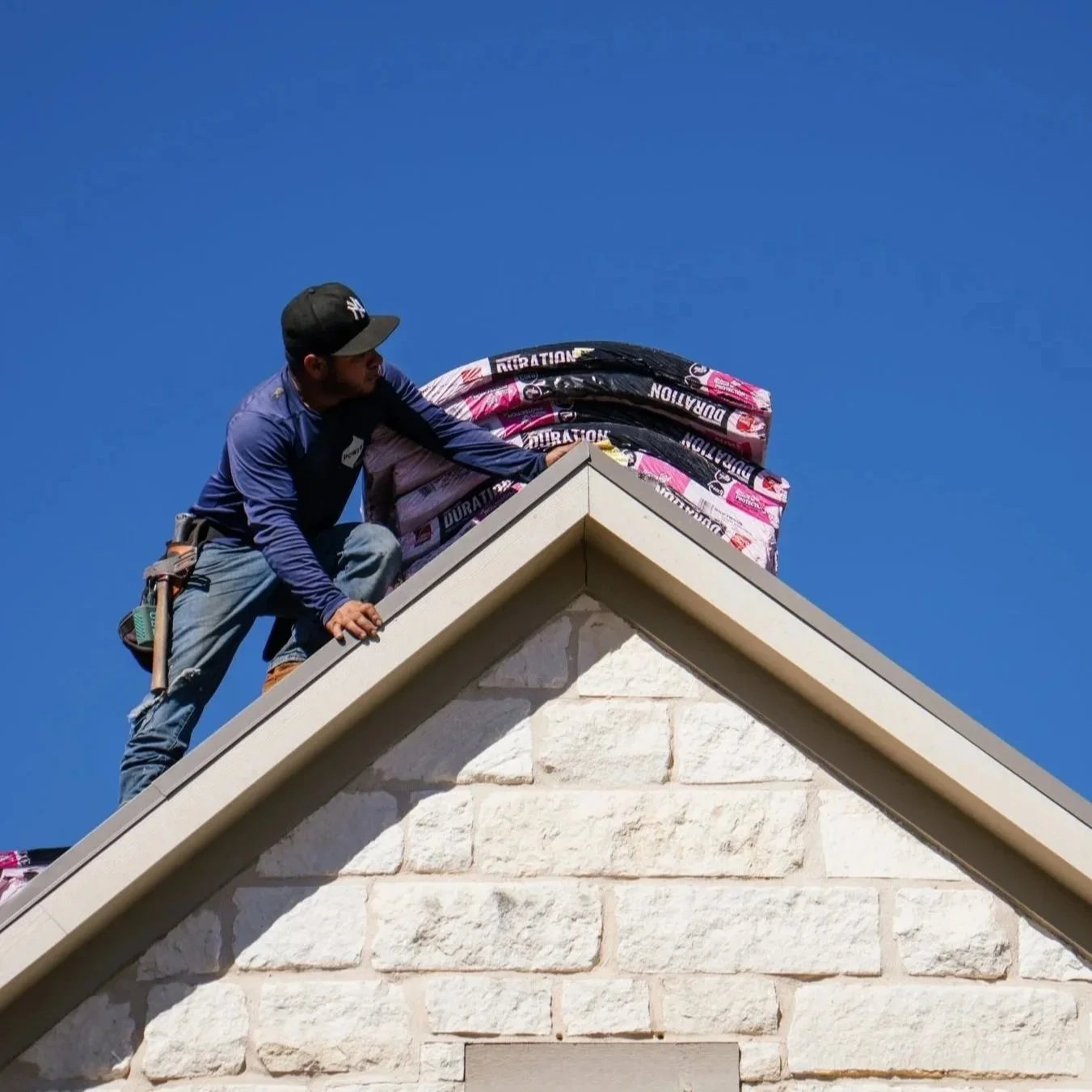 A man wearing a black cap, blue shirt, and jeans installing roofing materials on the corner of a building with stone exterior, under a clear blue sky.