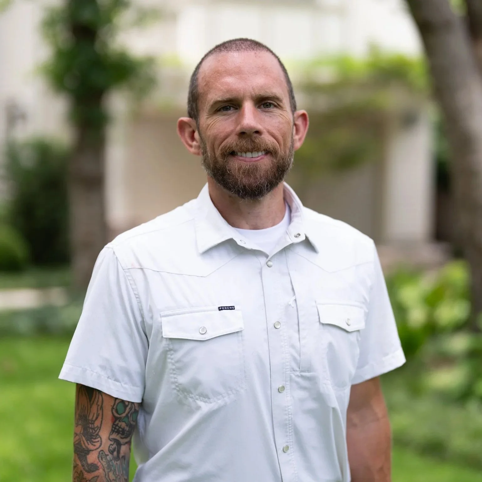 A man with a beard and closely cropped hair smiling outdoors, wearing a white button-up shirt with two breast pockets and a patch on the left pocket, standing in a green garden with trees and bushes in the background.