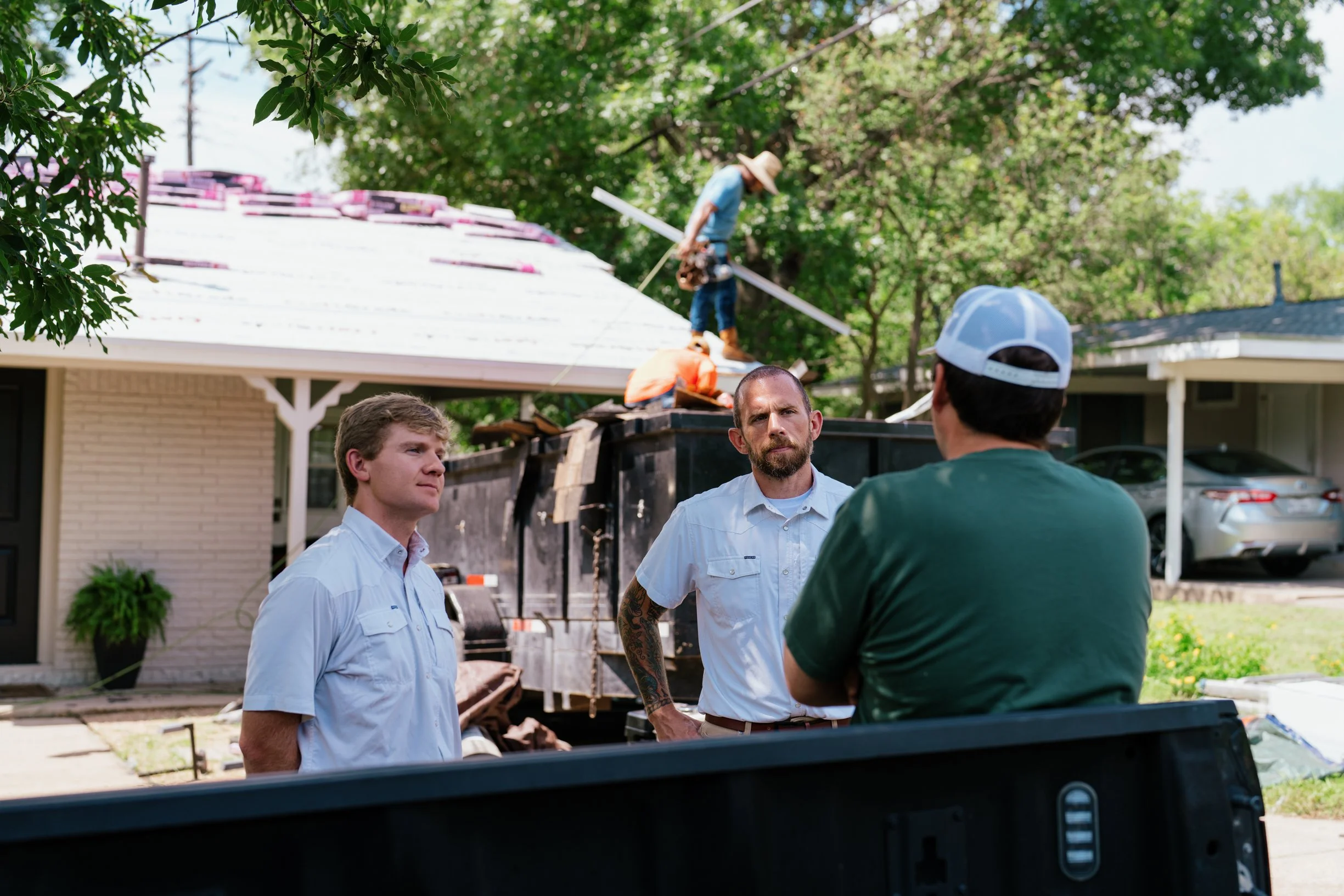 Three men stand in front of a vehicle, discussing a roofing project while a worker in the background installs new shingles on a house roof, under a tree and clear sky.