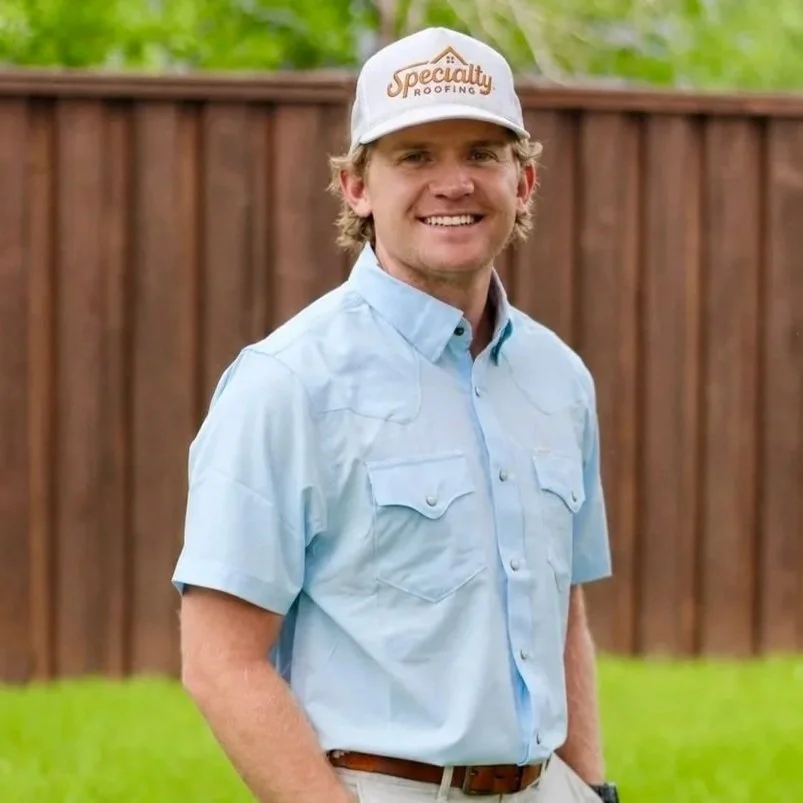 A young man smiling outdoors, wearing a light blue short-sleeved button-up shirt and a white baseball cap with 'Specialty Roofing' embroidered on it. Background includes a wooden fence and green trees.