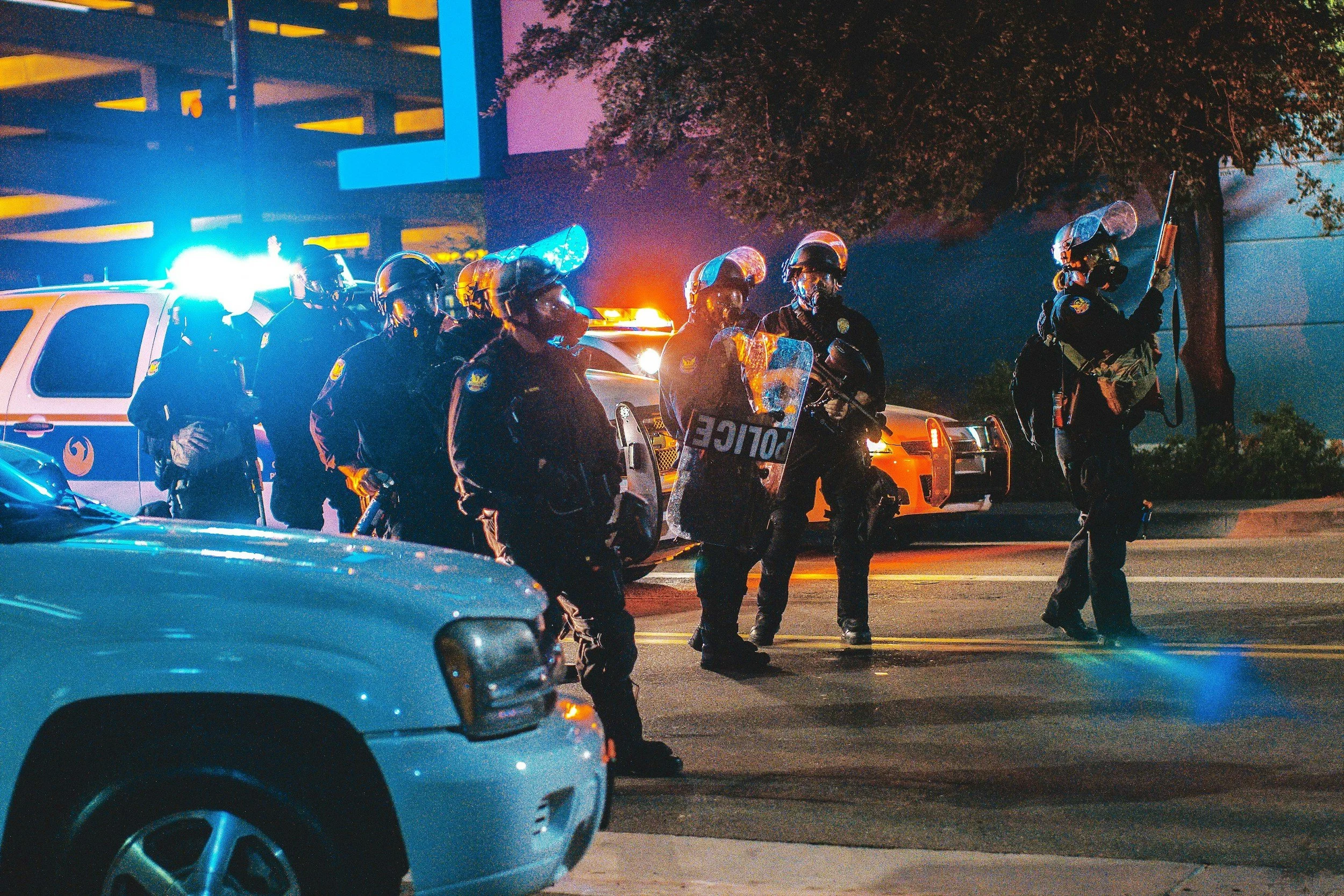 Nighttime scene of several police officers in riot gear, some holding shields and batons, standing on a city street with police cars with flashing lights in the background.