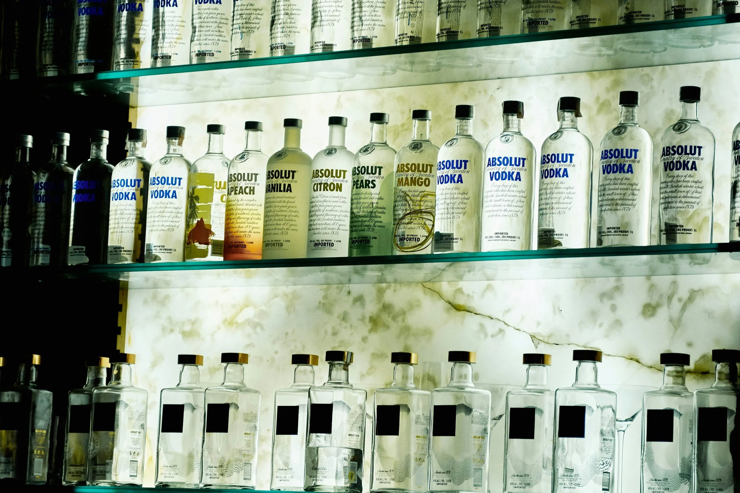 Display of various flavored vodka bottles on glass shelves in a store, including peach, vanilla, citroen, pear, and mango flavors.