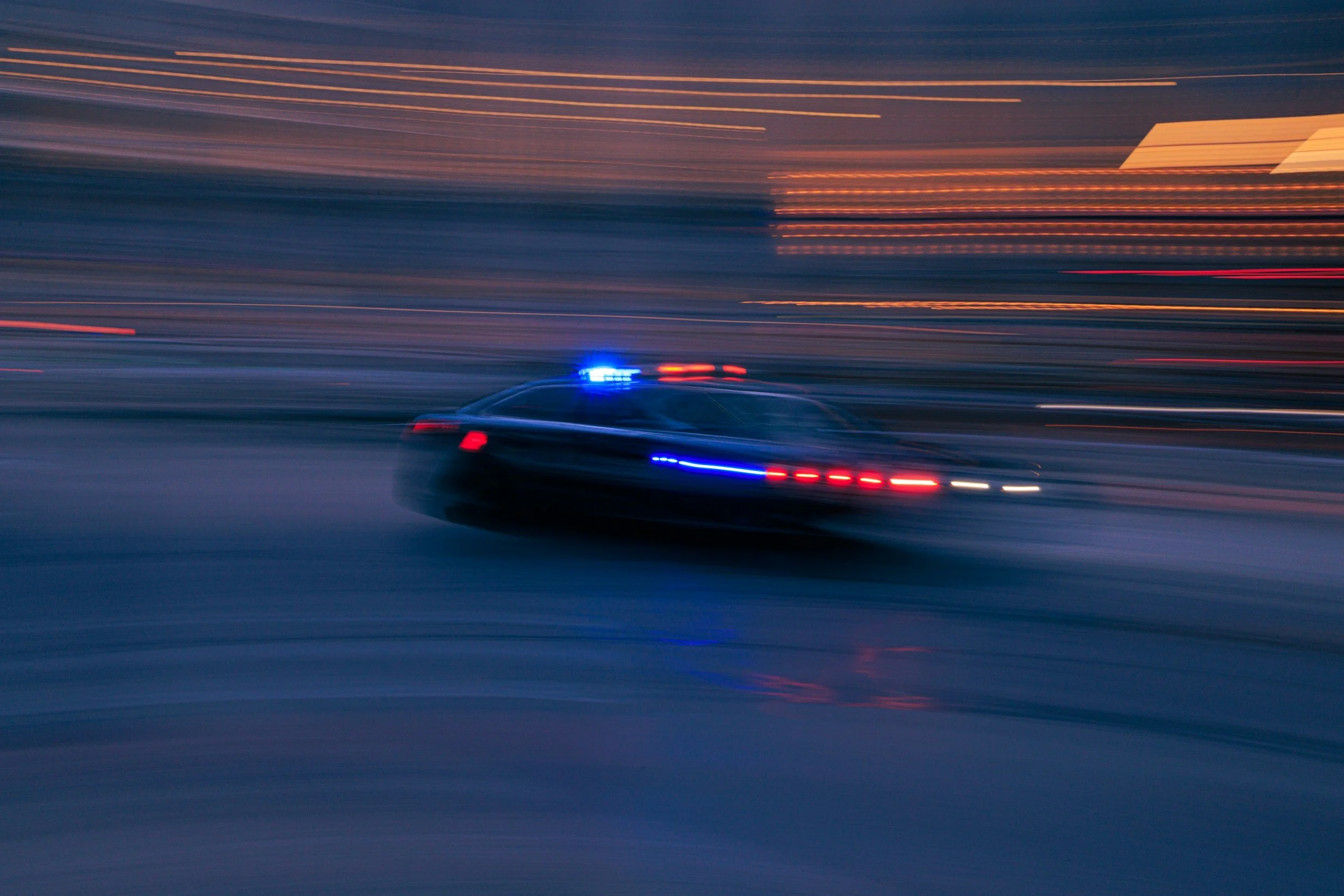 A police car with flashing blue and red lights speeding through a city street at night, with blurred lights in the background.