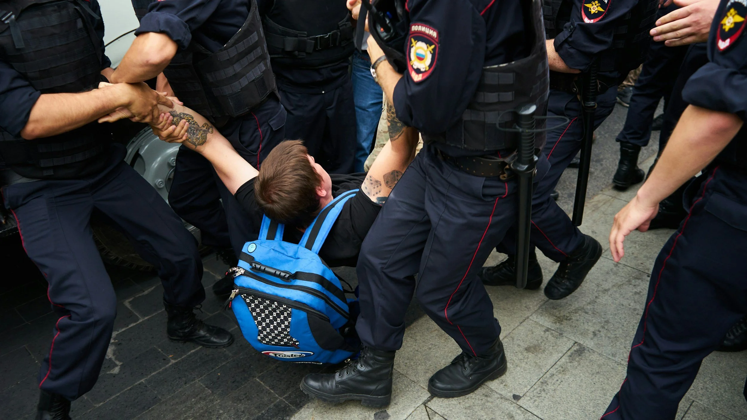 A man is being detained by multiple law enforcement officers on a city street. The man is on the ground, with his arm being held by officers, and is wearing a black shirt and a blue backpack. The officers are dressed in dark uniforms, some with badges and patches, and are surrounding the man.