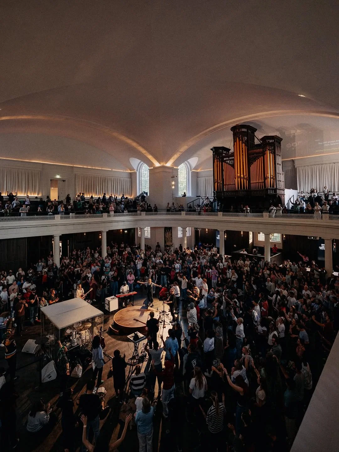 A large indoor concert hall filled with a crowd of people, with a stage and musicians performing beneath and above a pipe organ.