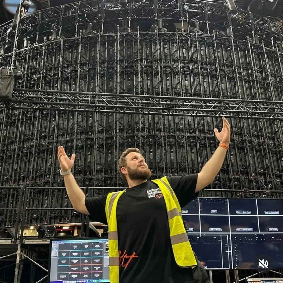 A man with a beard and wearing a black t-shirt and a yellow safety vest stands with arms raised in front of a large, complex stage rigging structure at an event. The background shows multiple screens and computer monitors.