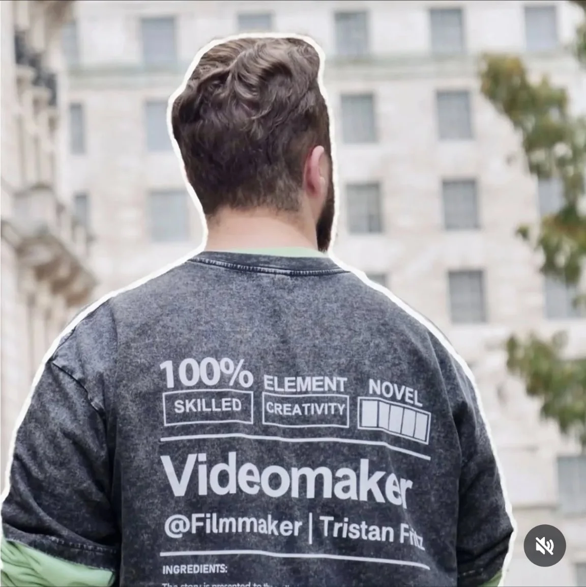 Back view of a man wearing a dark gray shirt with white text and graphics, standing outdoors with a building and trees in the background.