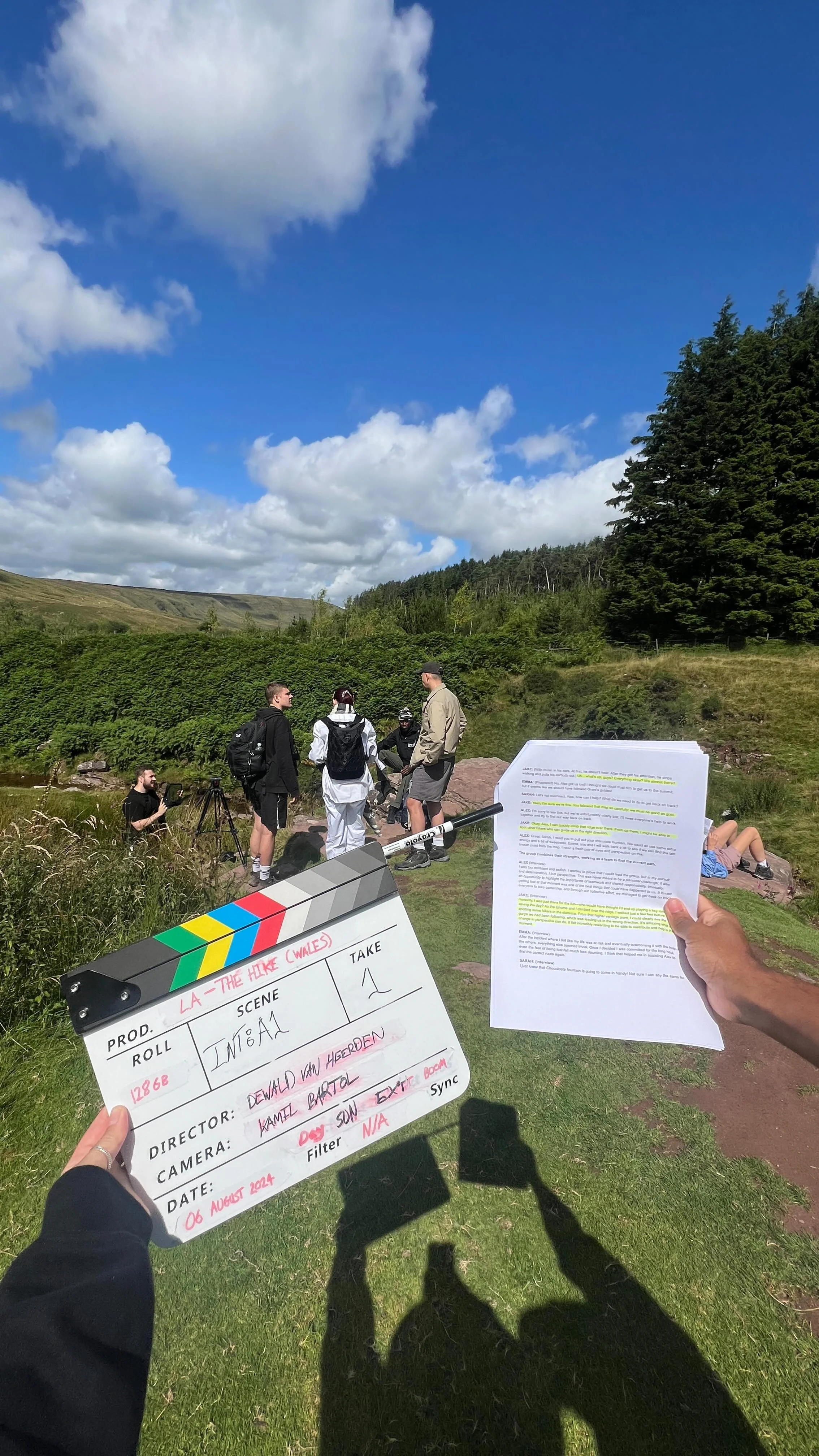 Film crew setting up for a shoot outdoors on a grassy field with a group of people standing and talking, surrounded by trees and hills under a partly cloudy sky.