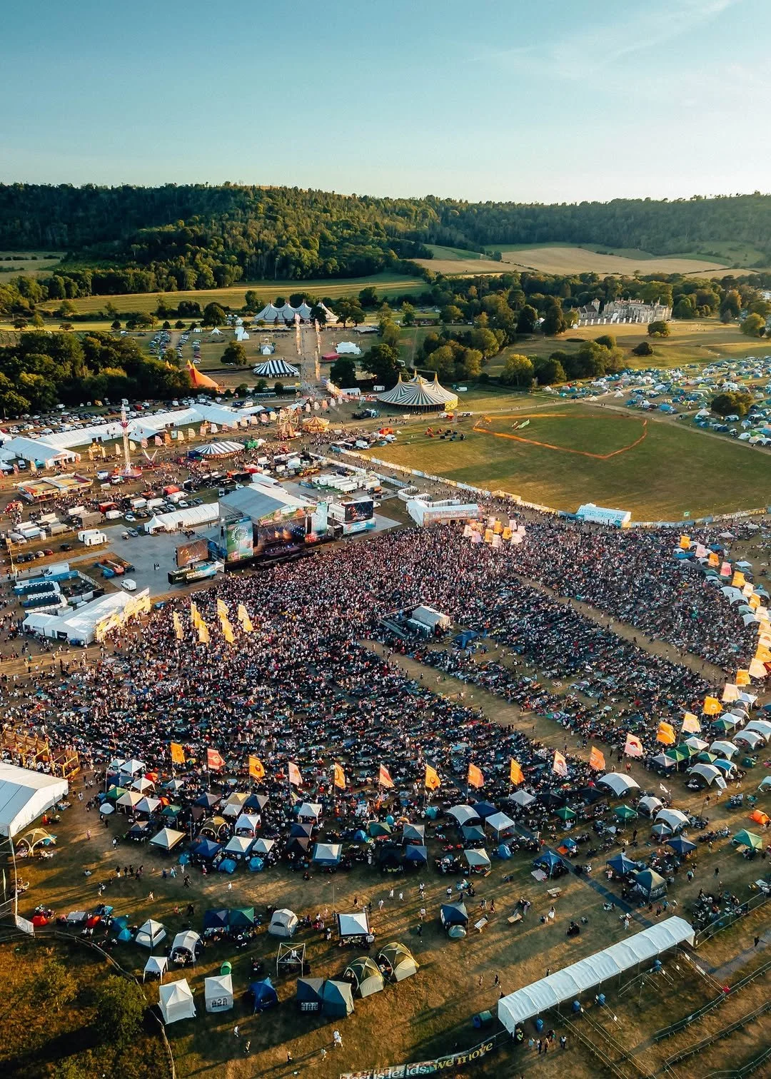 A large outdoor music festival with a crowd watching a stage, surrounded by tents, food stands, and amusement rides, set in a rural area with rolling hills and trees.