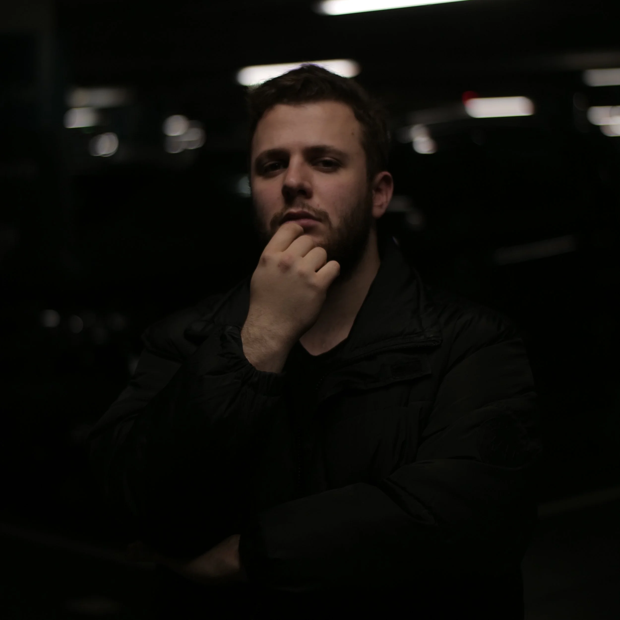 Tristan Fritz. A young man with a beard and short hair, wearing a black jacket, posing thoughtfully in a dimly lit environment with blurred lights in the background.