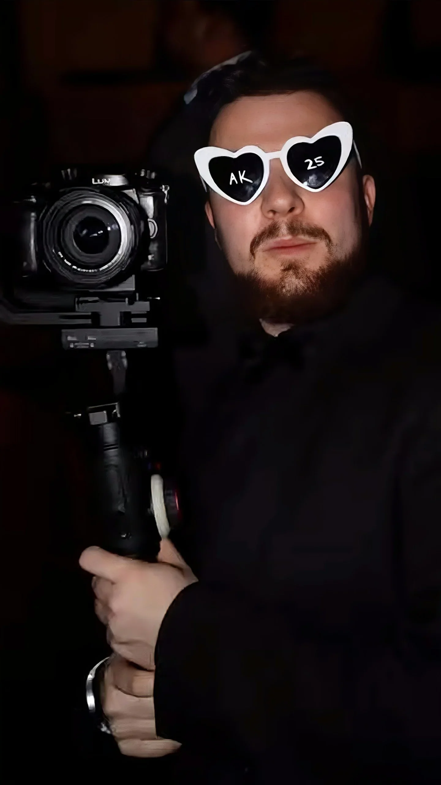 A man with a beard and mustache wearing sunglasses with heart-shaped frames that have 'AK' and '25' on the lenses, holding a professional camera on a stabilizer, taking a selfie or photo.