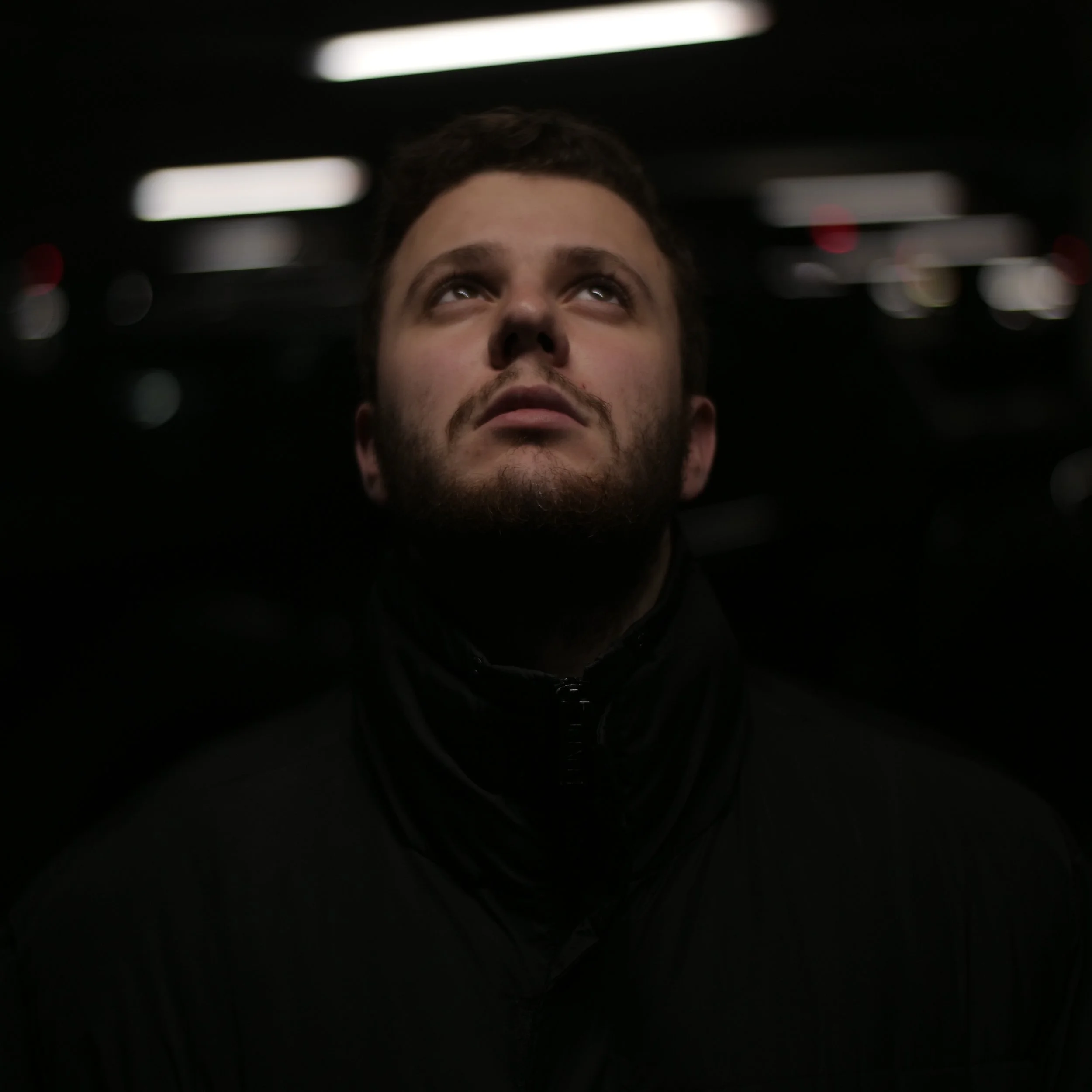 Tristan Fritz. A man with a beard looking upward in a dark indoor setting with blurred lights in the background.