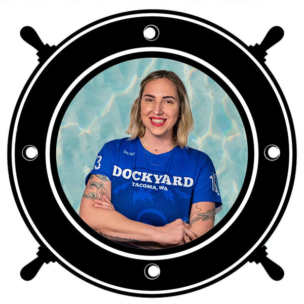 A woman with blonde hair and tattoos, smiling and holding a gold helmet, seen through a circular porthole. 