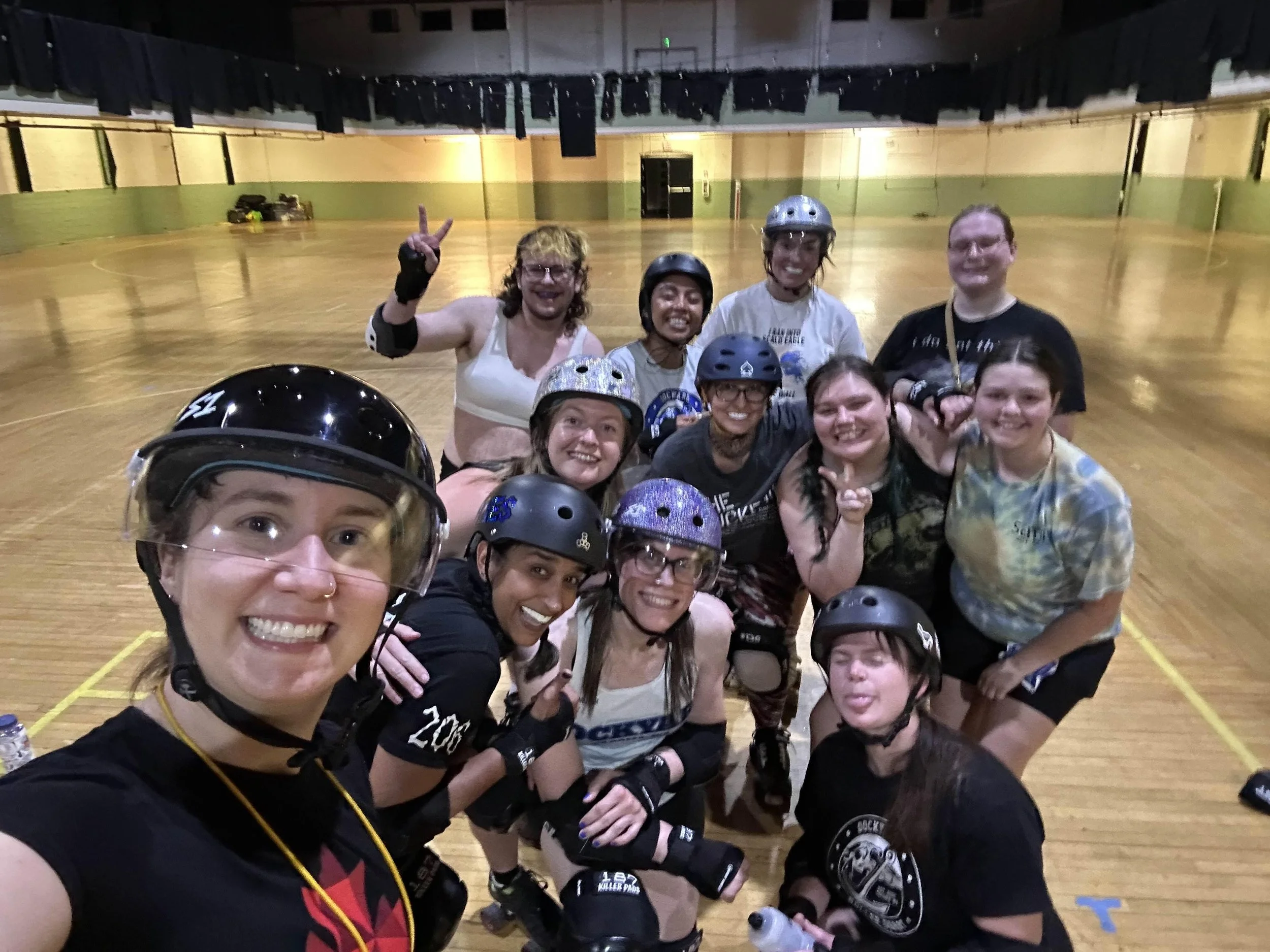 A group of people in roller derby gear, including helmets and protective pads, smiling and posing together inside a roller skating rink.