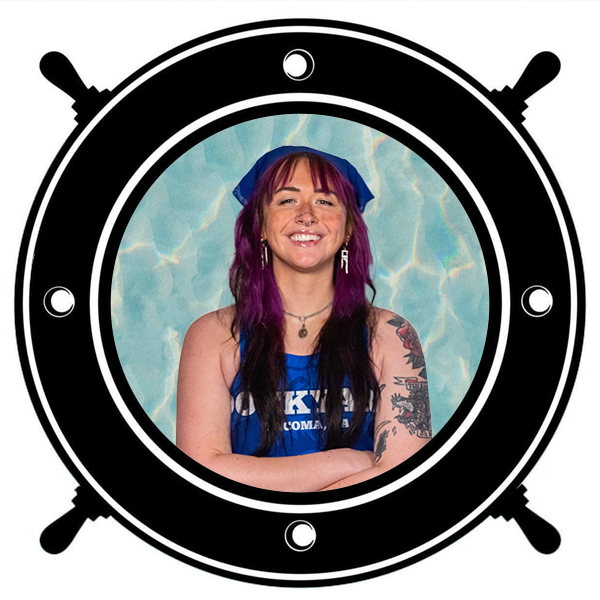 A young woman with purple and black hair smiling and posing with her hands under her chin, seen through a circular porthole.