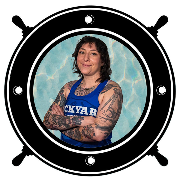 Woman with tattoos wearing a blue tank top holding a white helmet with colorful designs, seen through a circular porthole. 