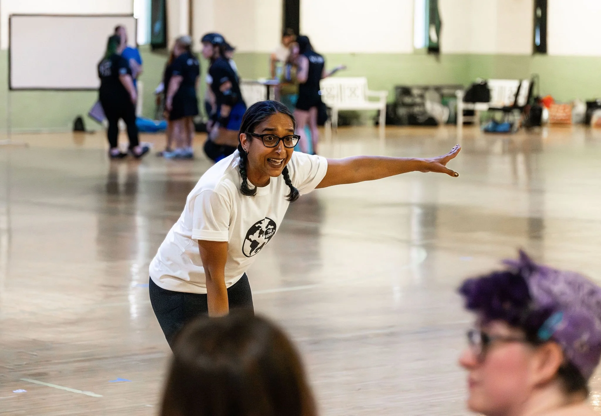 A woman with glasses and braids, wearing a white T-shirt with a globe graphic, is pointing to the right while speaking to a group of people in a large, indoor space with a wooden floor. In the background, several women are standing and talking.