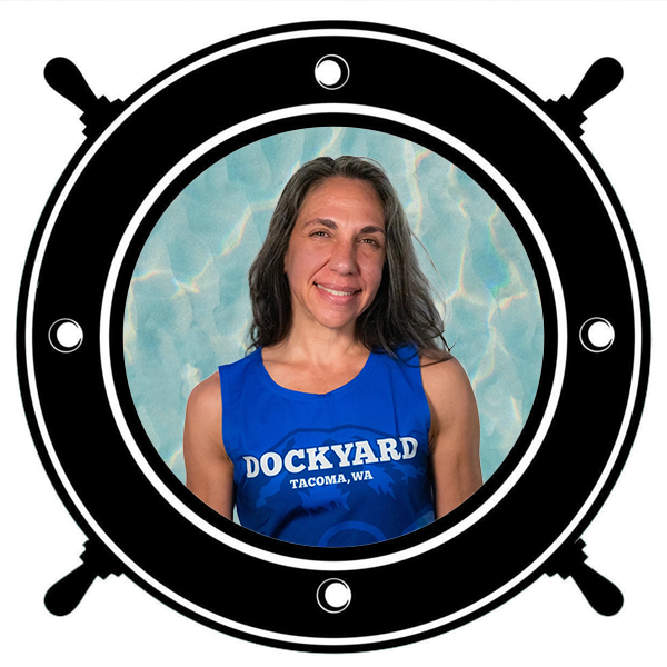 A woman in a blue tank top with the word "Dockyard" and "Tacoma, WA" printed on it, smiling with her arms crossed, seen through a circular porthole. 