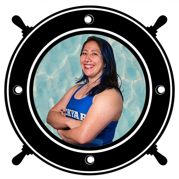 Young woman smiling in a swimming pool with long dark hair and blue streaks, wearing a blue tank top, seen through a circular porthole. 