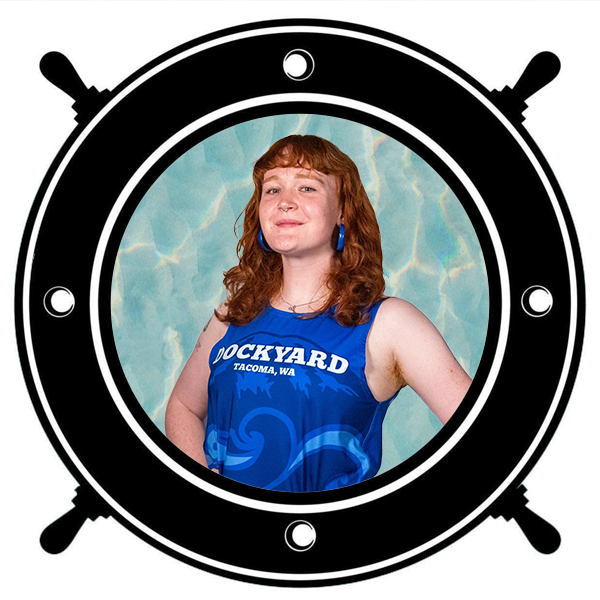 Young woman with red hair wearing a blue sports jersey with "Dockyard Tacoma, WA" written on it, holding a helmet labeled "Spikes" on top seen through a circular porthole.