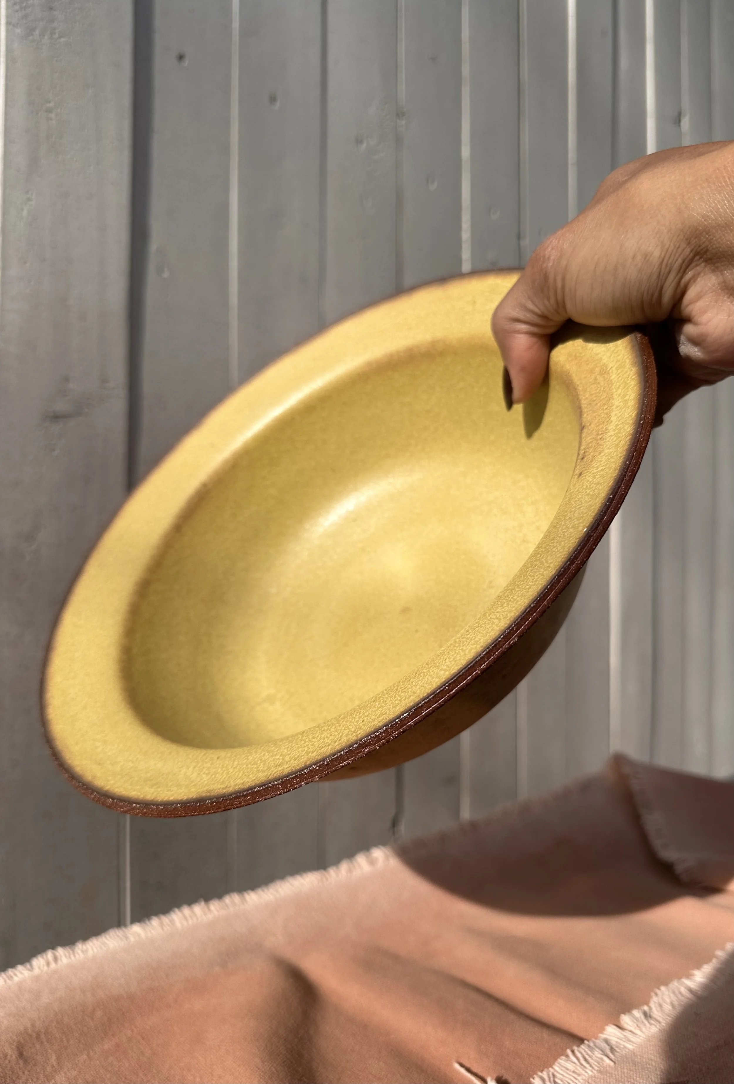 A person holding a yellow ceramic bowl with a brown exterior, against a wooden wall background.