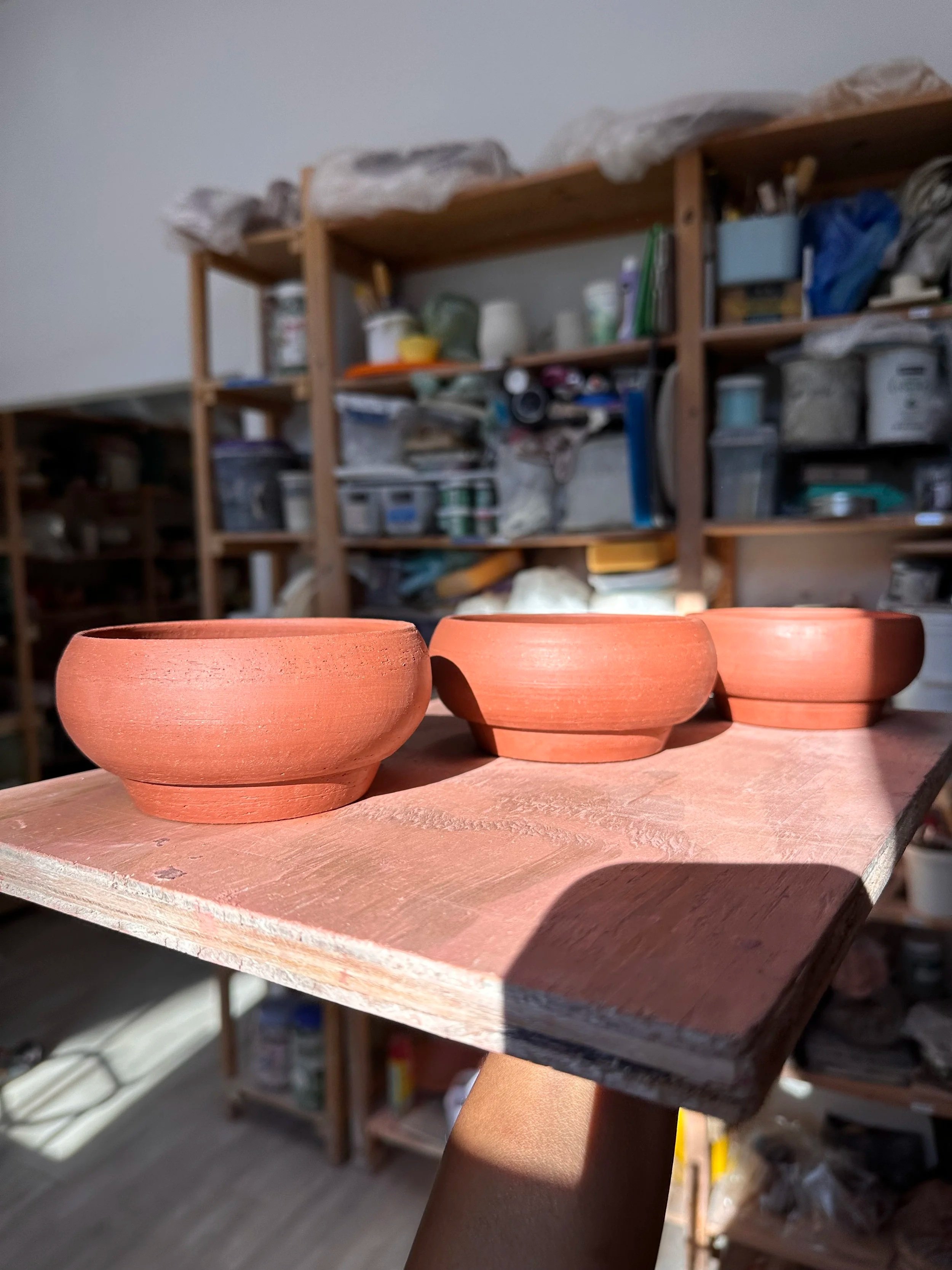 Three unfinished terracotta bowls on a wooden shelf in a pottery studio.