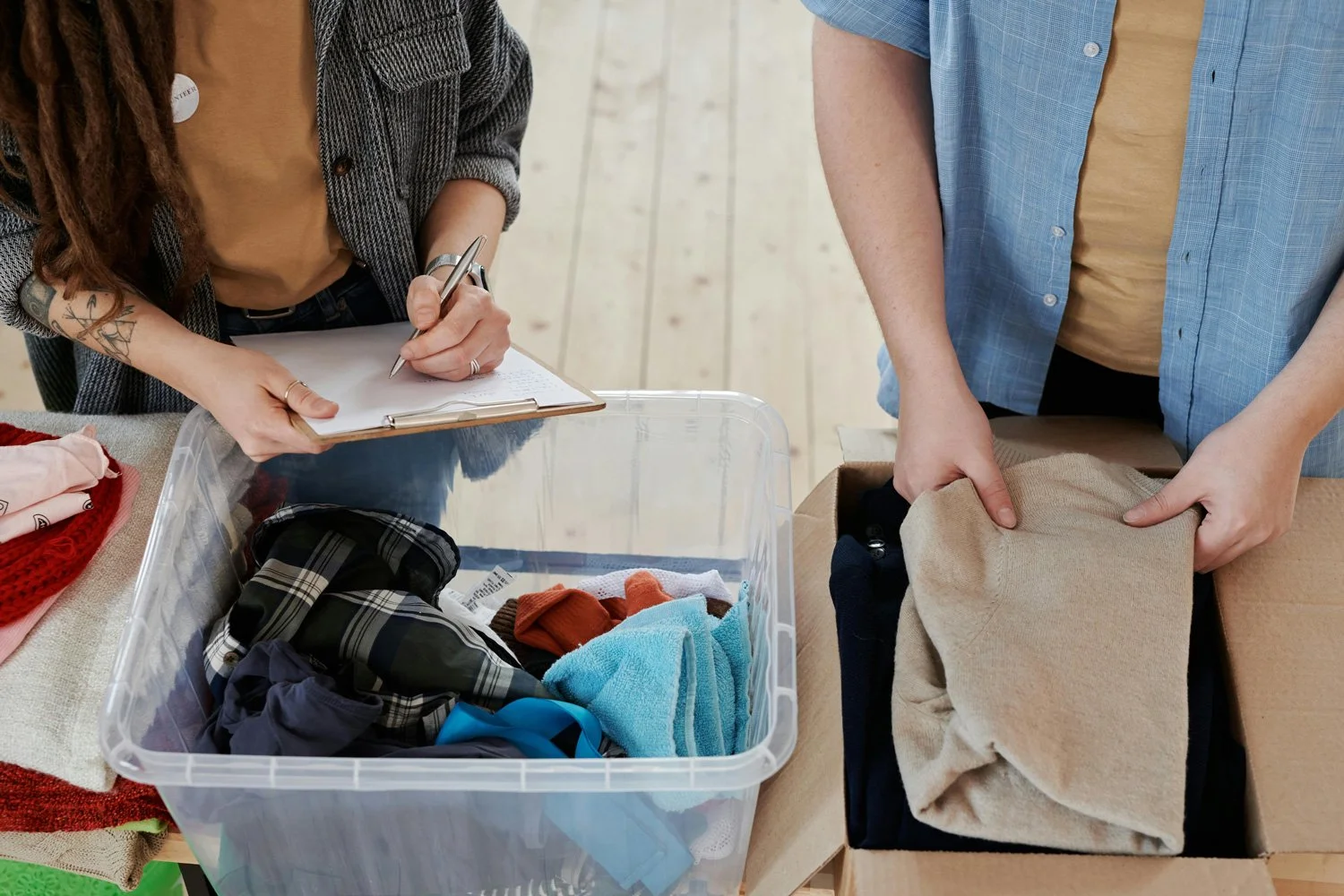 Two people are sorting clothes, one with a clipboard and pen, the other holding folded clothes over a cardboard box, on a wooden table.