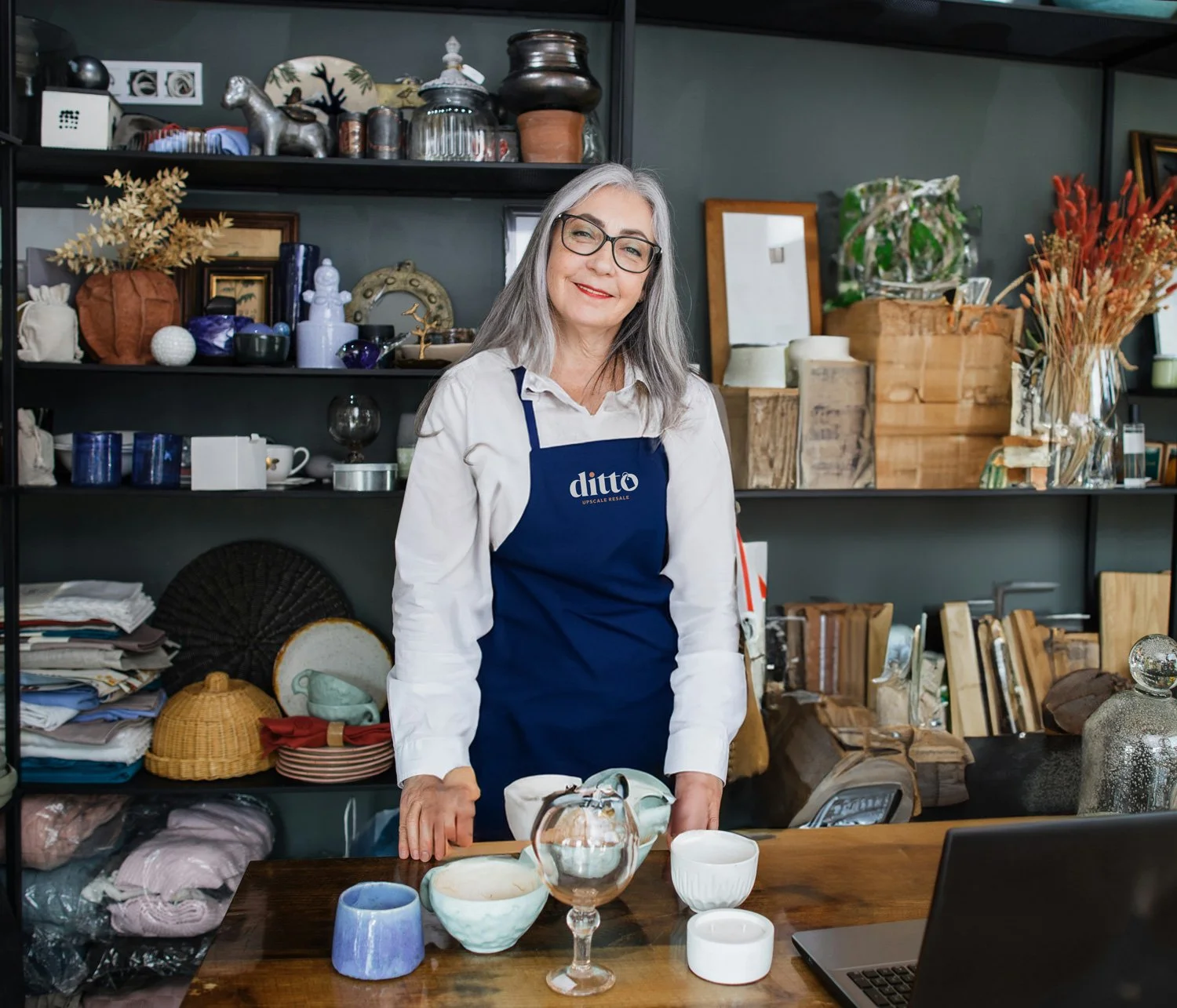 A woman in glasses and a blue apron stands behind a wooden table with various ceramic and glass items, smiling at the camera. Shelves behind her hold decorative objects, jars, books, and dried flowers.