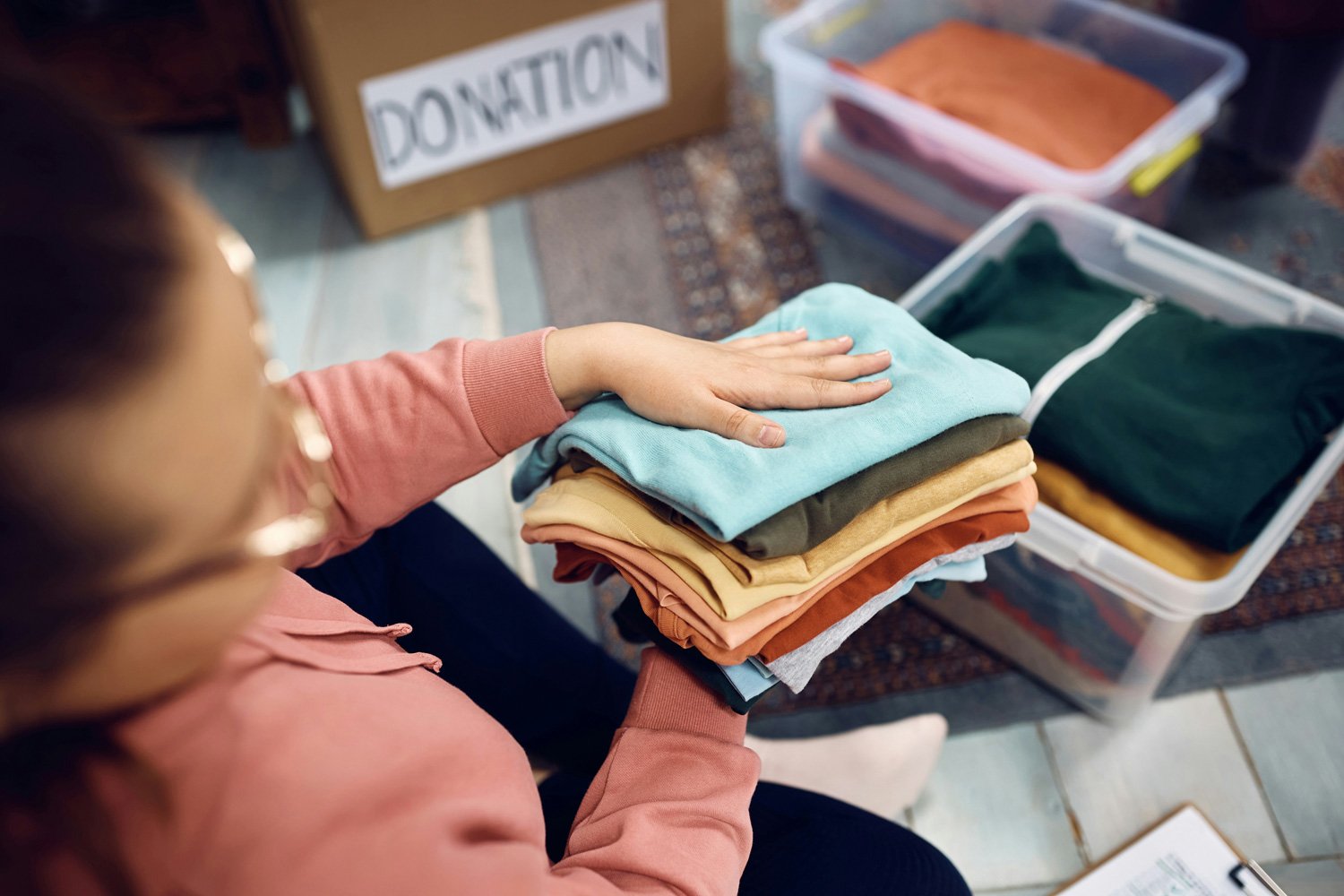 A person sorting folded clothes, including various colored shirts, in a donation setup with a box labeled 'DONATION' and plastic containers filled with clothes.