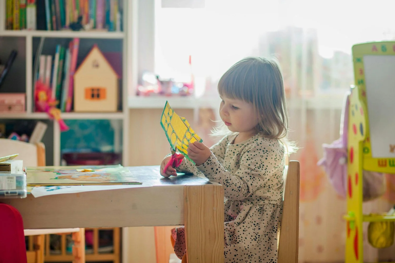 A young girl with shoulder-length hair sitting at a wooden table, using pink scissors to cut yellow and green patterned paper in a brightly lit room with bookshelves and toys in the background.