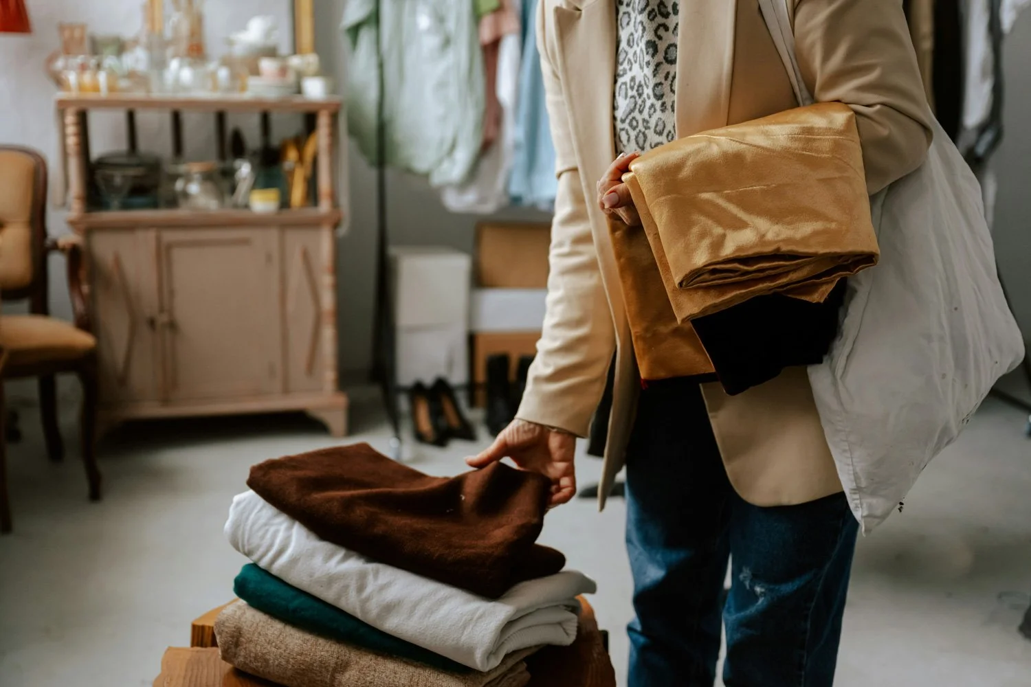 Person shopping for clothes, holding folded fabric or clothing items in a thrift or vintage shop, with a stack of clothes on a table in front.