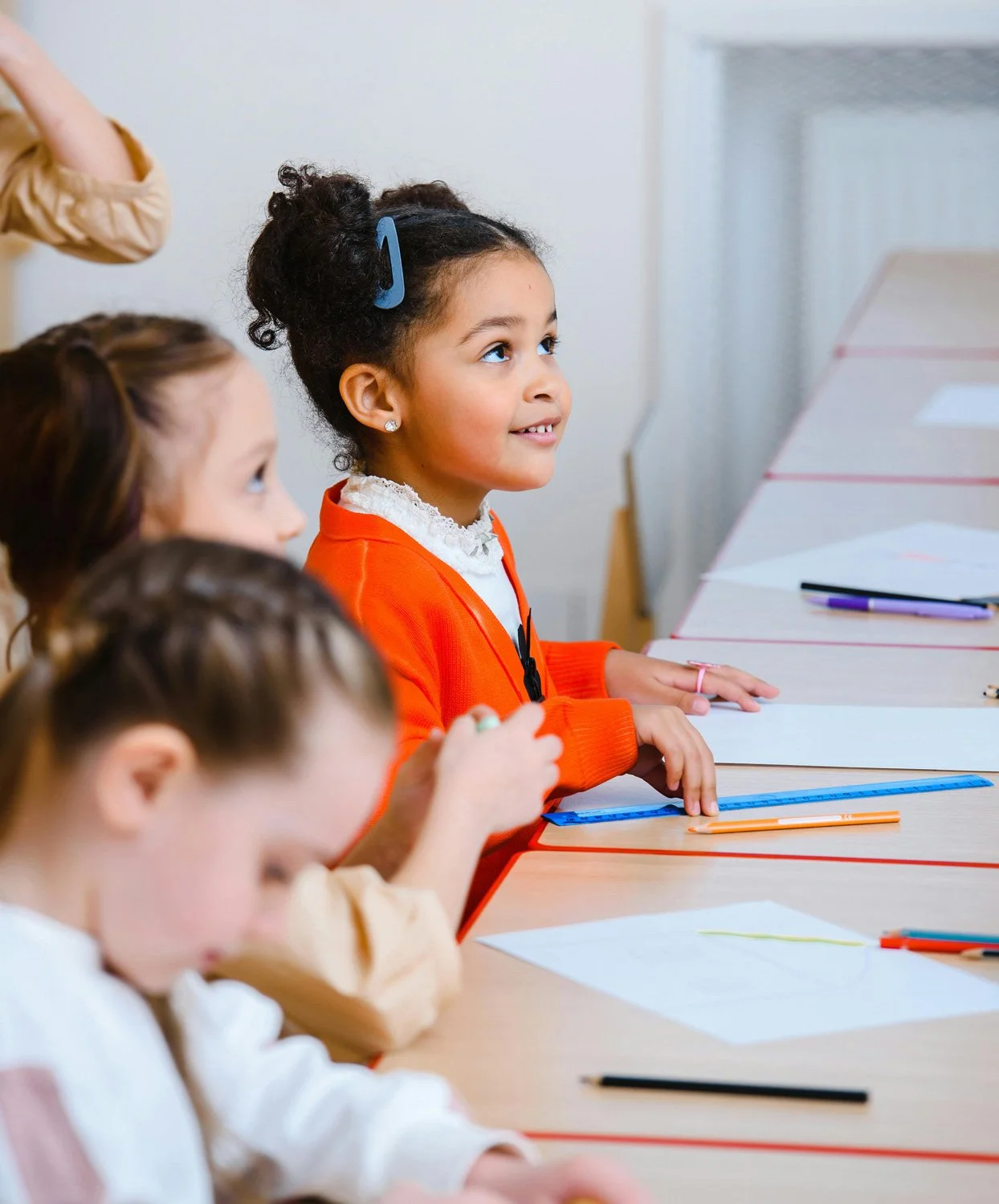 A group of young girls seated at desks in a classroom, with one girl in an orange sweater smiling and looking attentively at something off-camera.