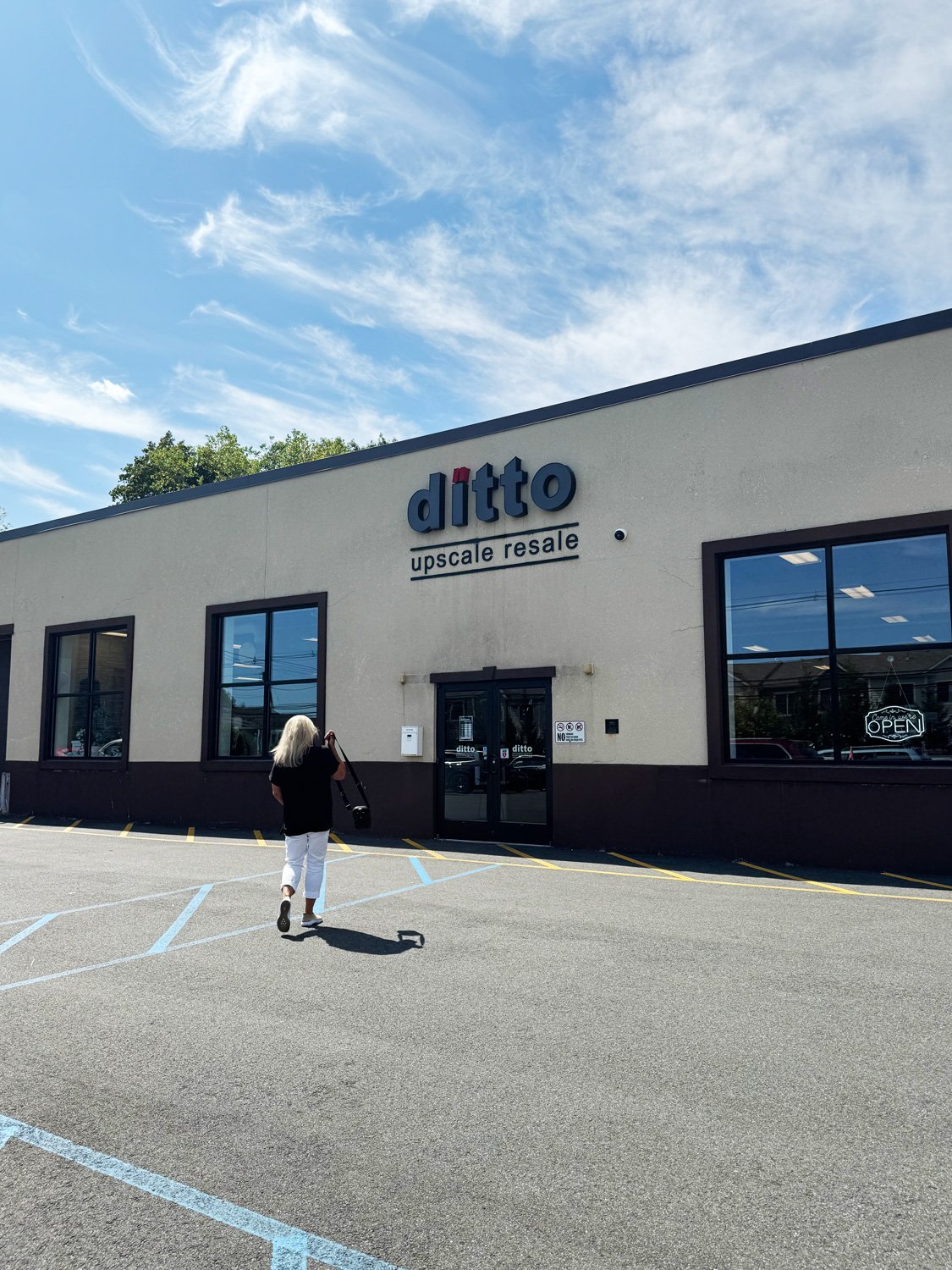 Front of a retail store named 'ditto' with a sign that reads 'upscale resale'. A woman with long white hair, wearing a black top and white pants, is walking toward the entrance. The parking lot has painted lines, and the sky is blue with some clouds.