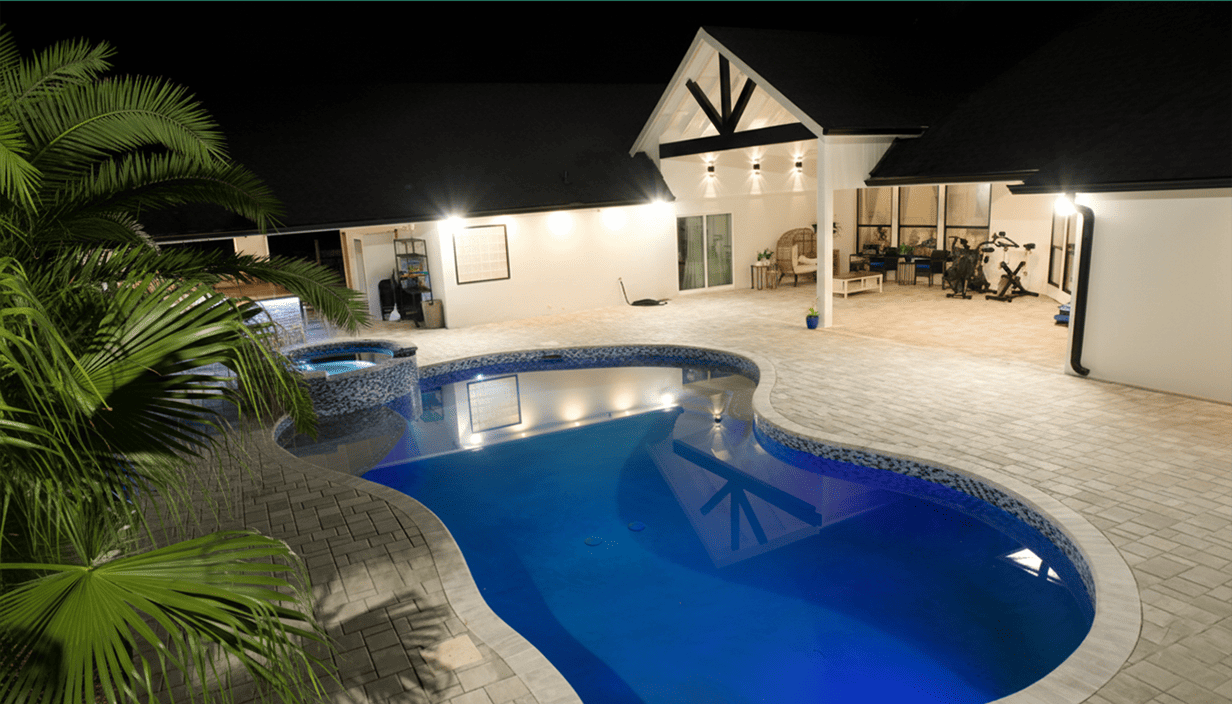 Nighttime backyard scene featuring a lit kidney-shaped swimming pool with underwater lights, surrounded by pavers, with a nearby palm tree, outdoor seating area, exercise equipment, and a white house with black roofing and outdoor lighting.
