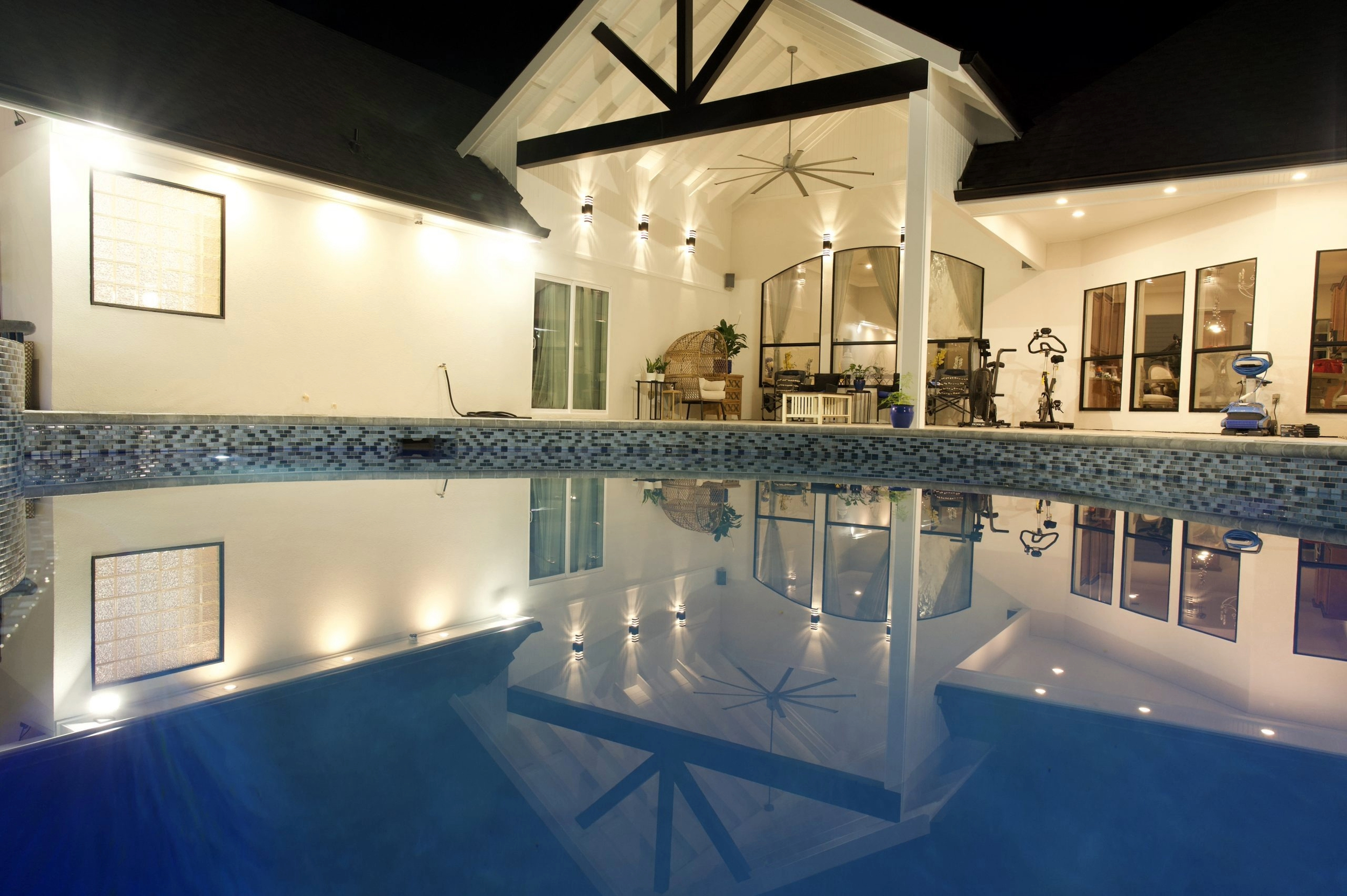 Nighttime view of an illuminated backyard with a swimming pool, covered patio with ceiling fan, workout equipment, and outdoor furniture reflected in the pool water.