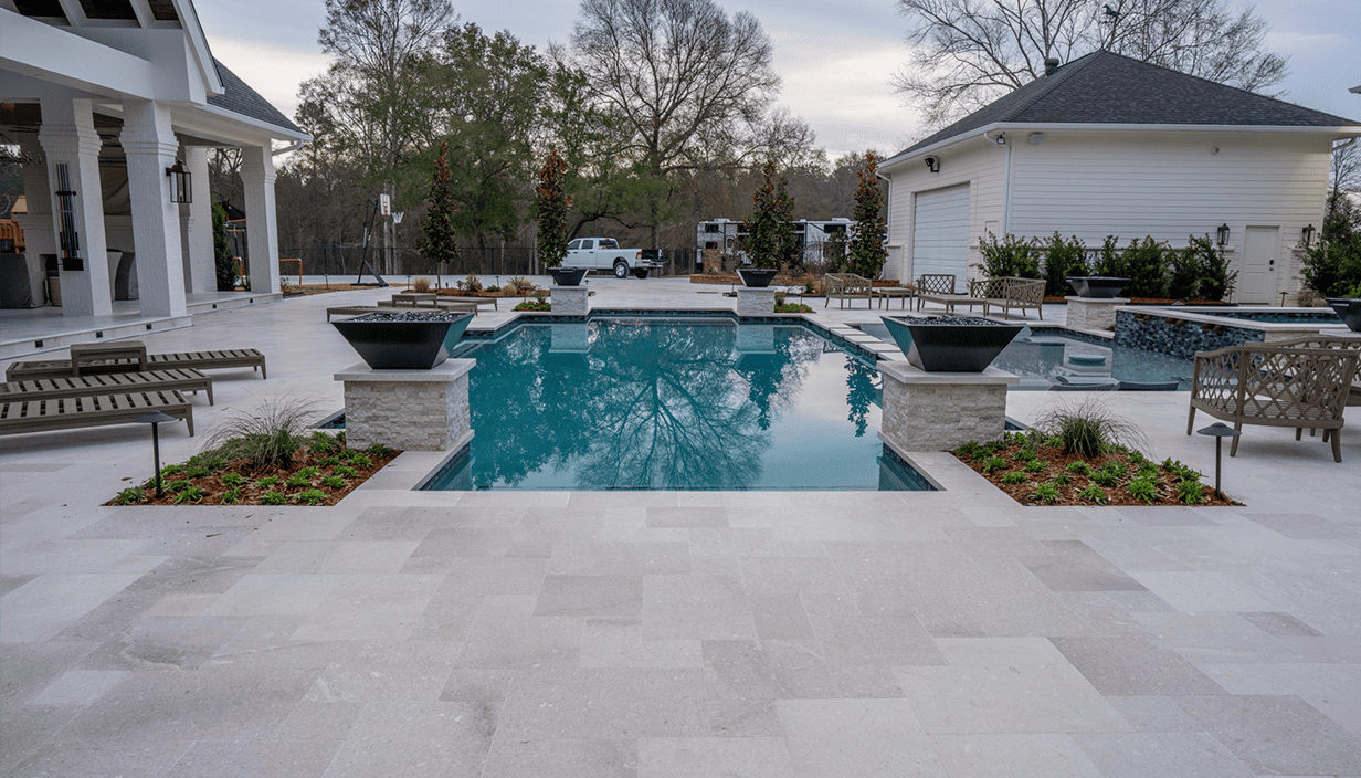 A modern backyard with a rectangular swimming pool, surrounded by light-colored stone decking, with lounge chairs, planters, trees, and a white house with a garage in the background.