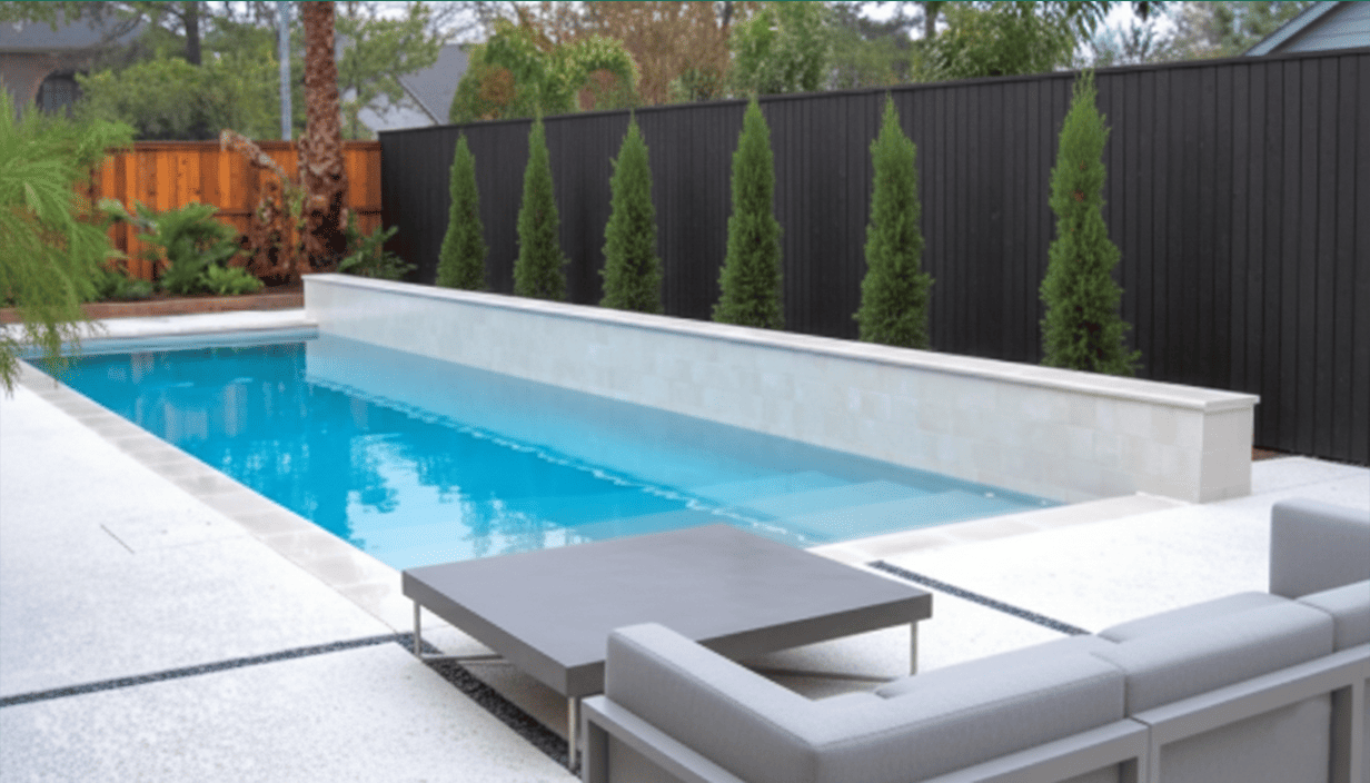 Modern backyard with a rectangular in-ground swimming pool, surrounded by white tile, with a gray table and seating area nearby. Tall, narrow evergreen trees line the black fence in the background.