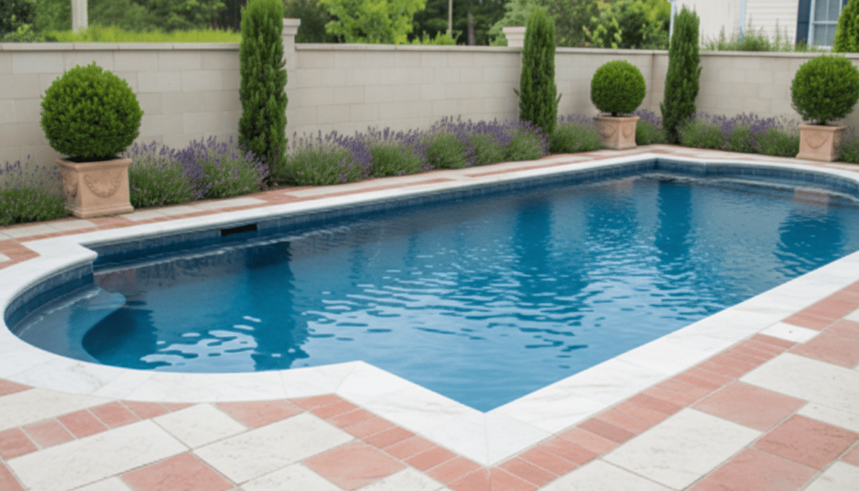 A rectangular swimming pool with a curved corner, surrounded by a brick and white stone patio, with a white wall and neatly trimmed bushes and trees in the background.